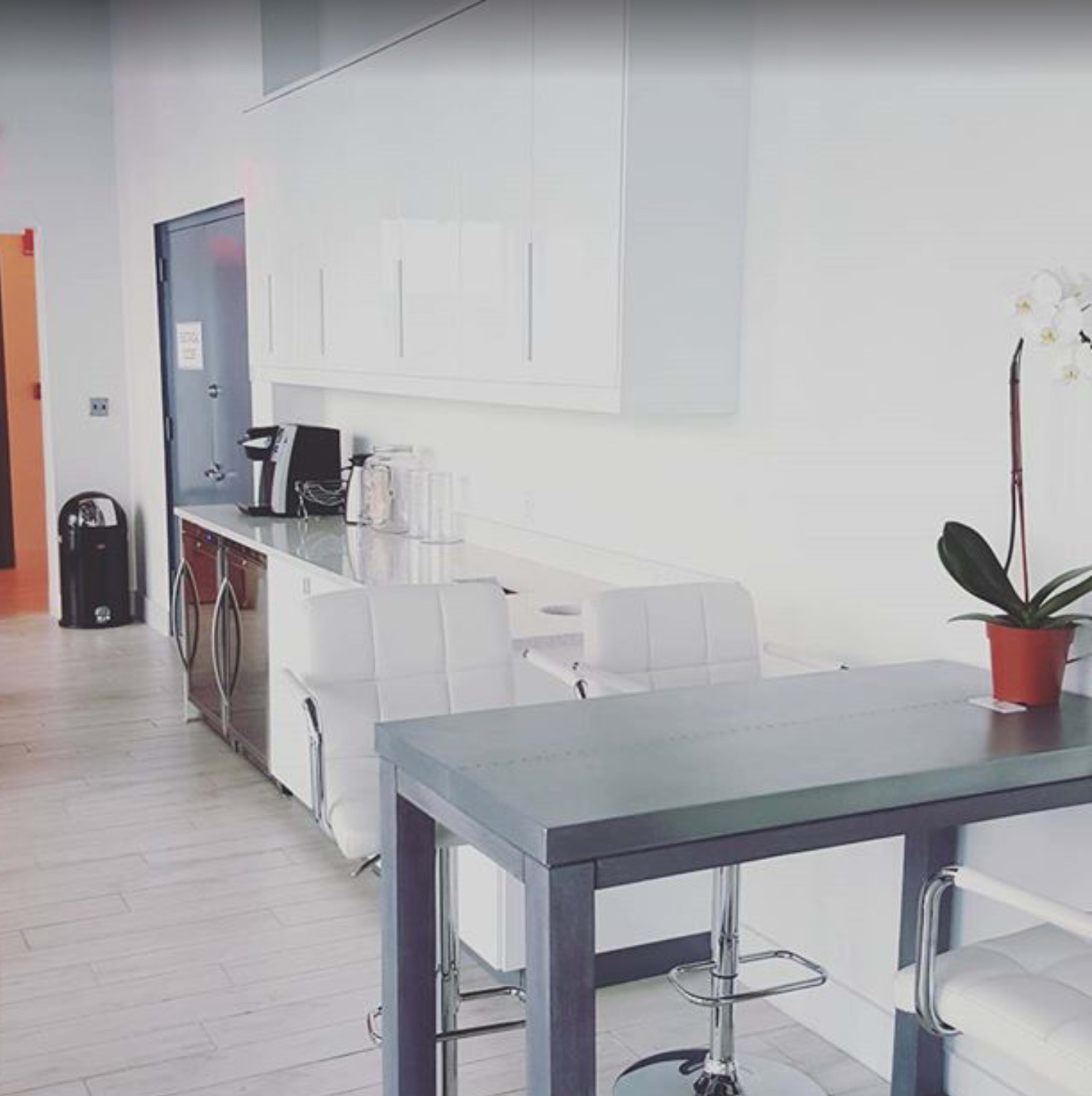 The image shows a modern kitchen area with white cabinetry, a coffee station, and a small dining table with bar stools.