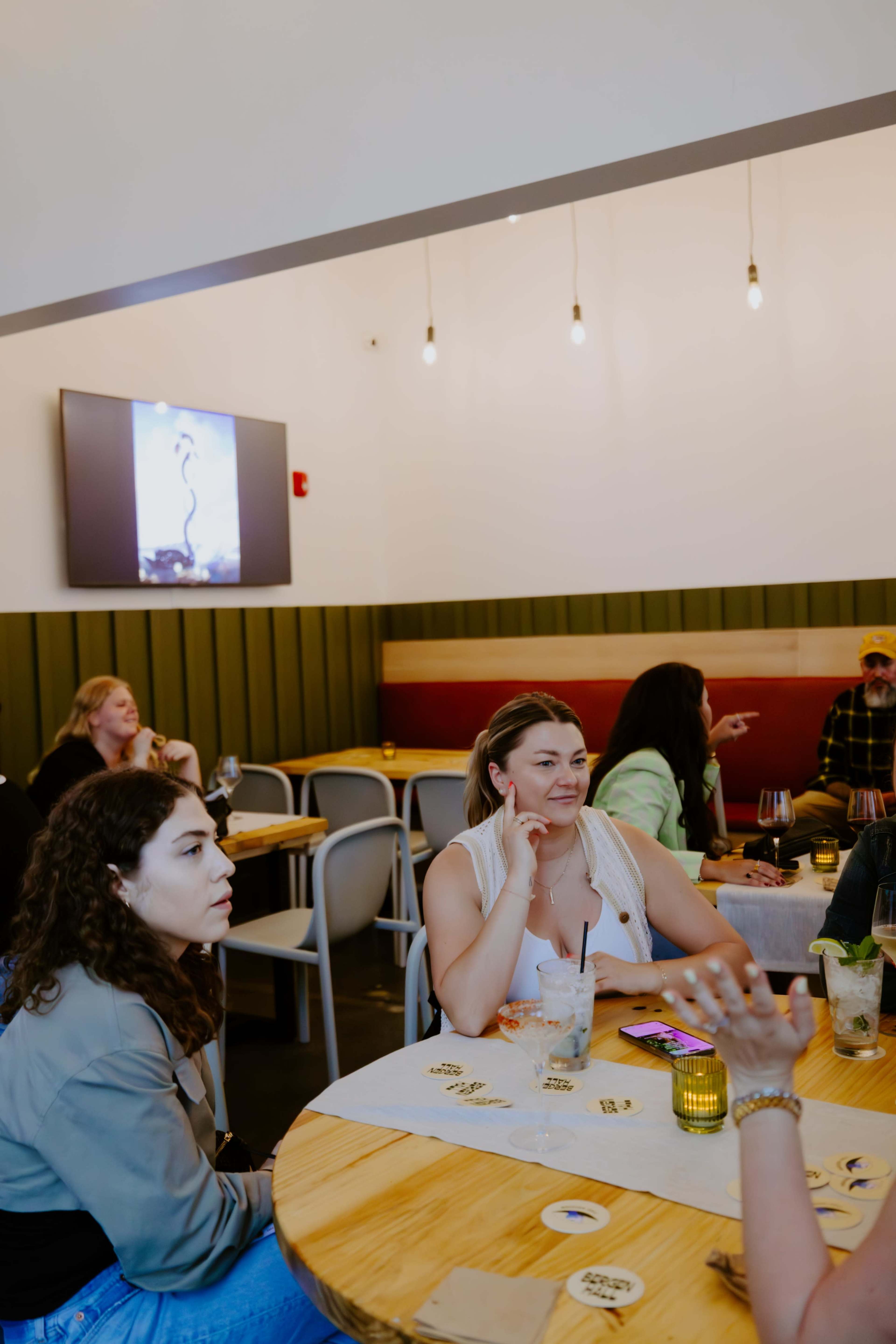 A group of people is seated at tables in a casual dining space, with some engaging in conversation and a large screen displaying an image on the wall.