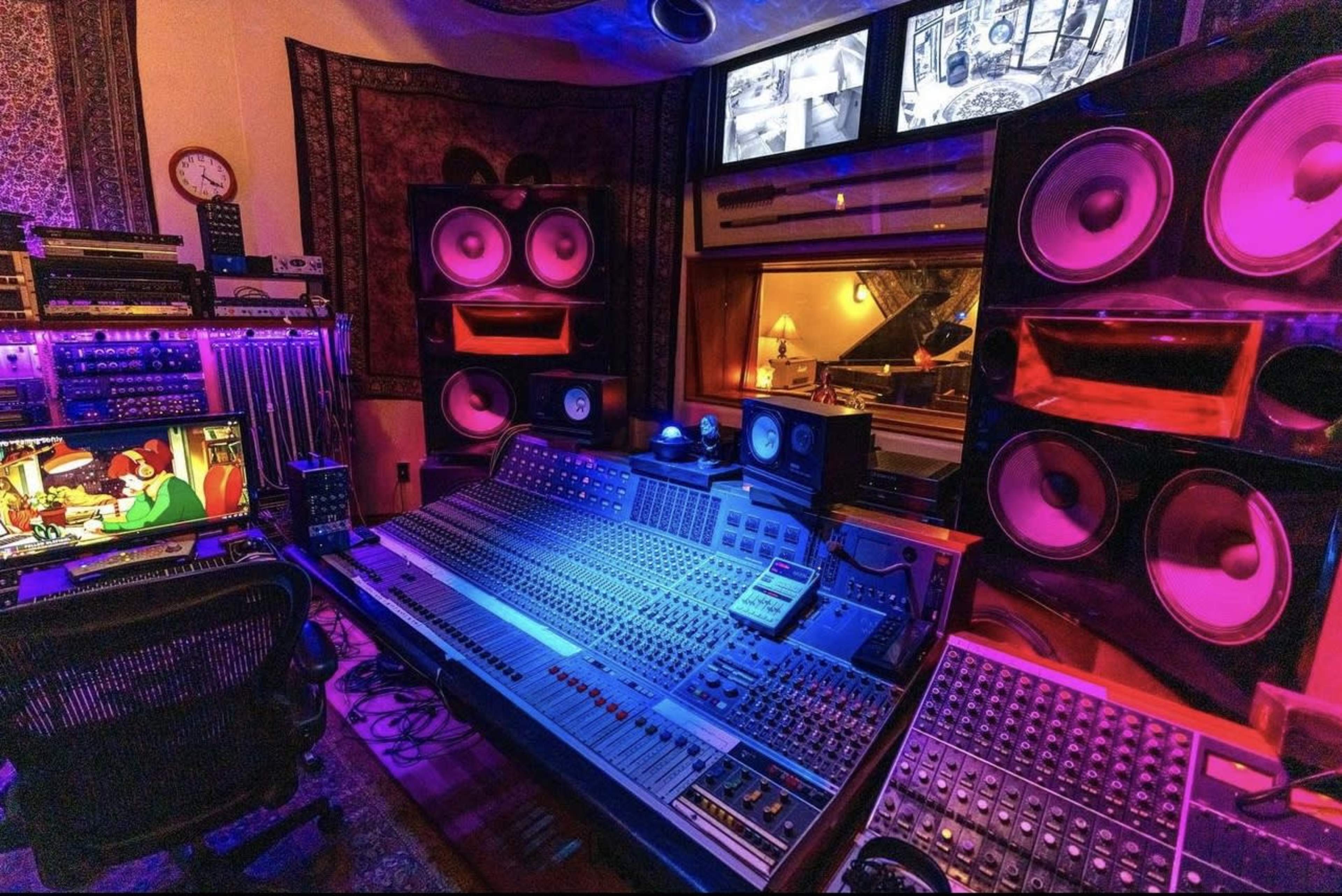 A music recording studio equipped with an extensive audio mixing console, large speakers, and multiple screens displaying various visuals.