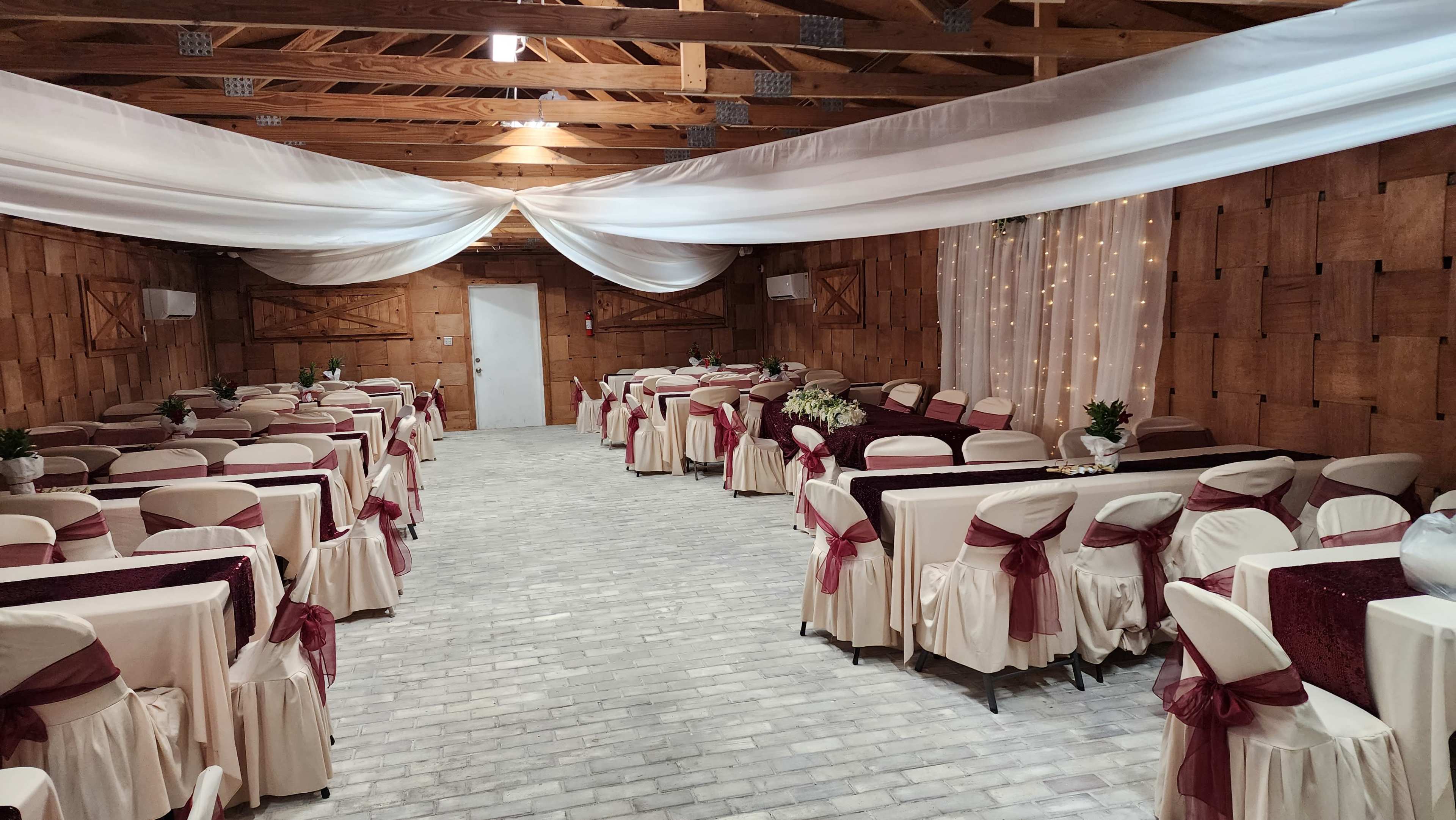 A banquet hall set up with rows of tables covered in beige tablecloths and burgundy sashes, under draped white fabric and soft lighting.