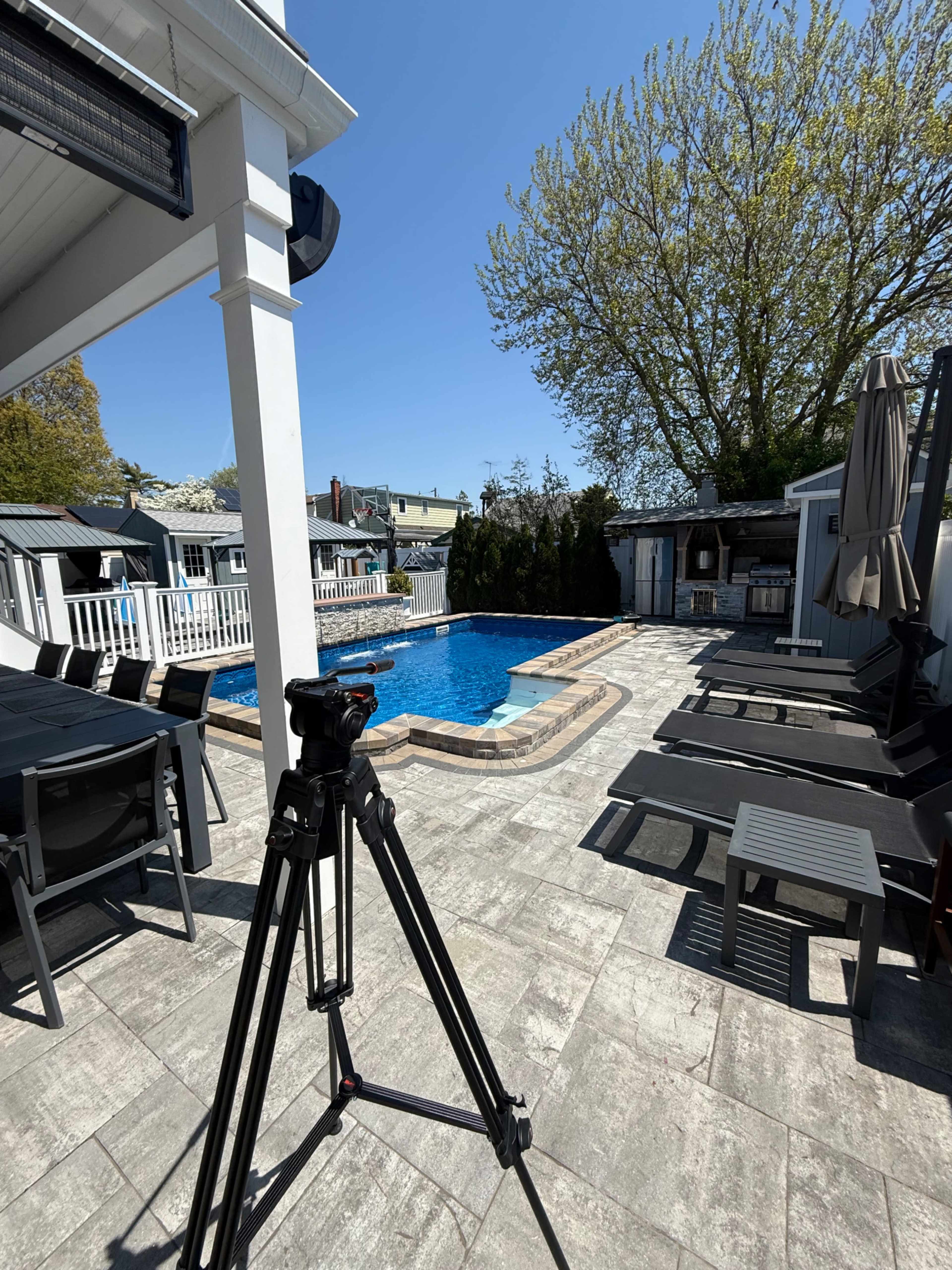 A tripod stands on a stone patio overlooking a swimming pool surrounded by lounge chairs and trees on a clear day.