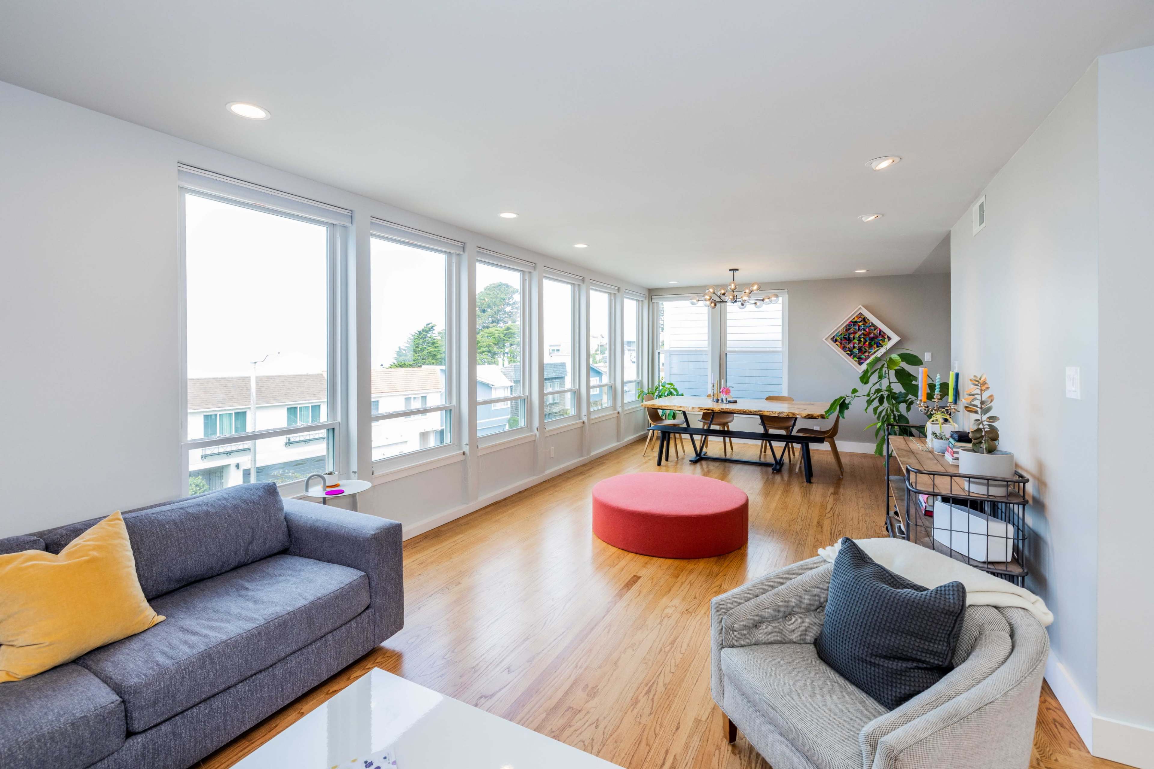 A spacious, brightly lit living room features large windows, modern furniture, and a dining area with a table and chairs.