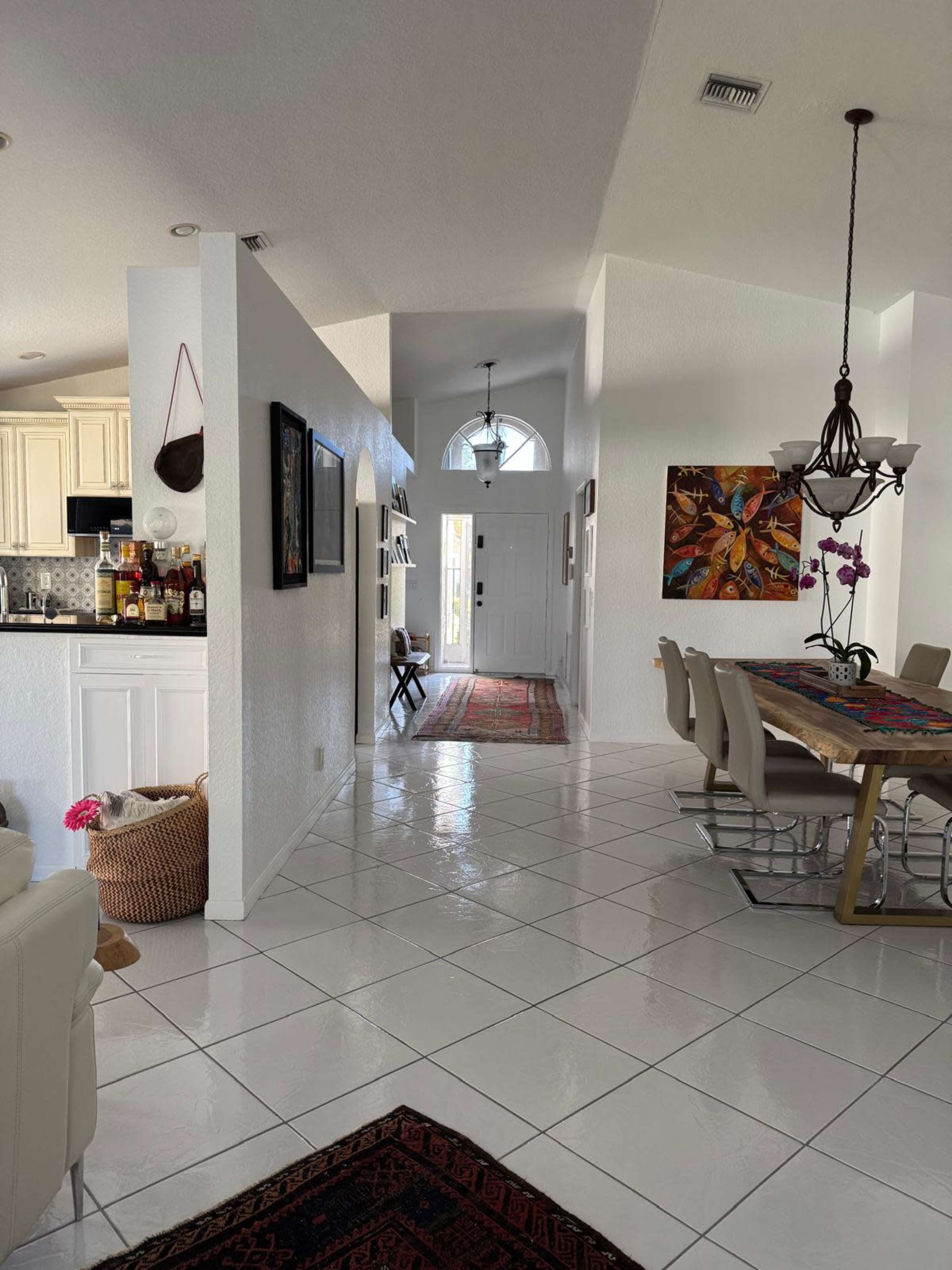 A spacious, brightly lit hallway leads from a kitchen area to a front door, with a dining table on one side and decorative art on the walls.
