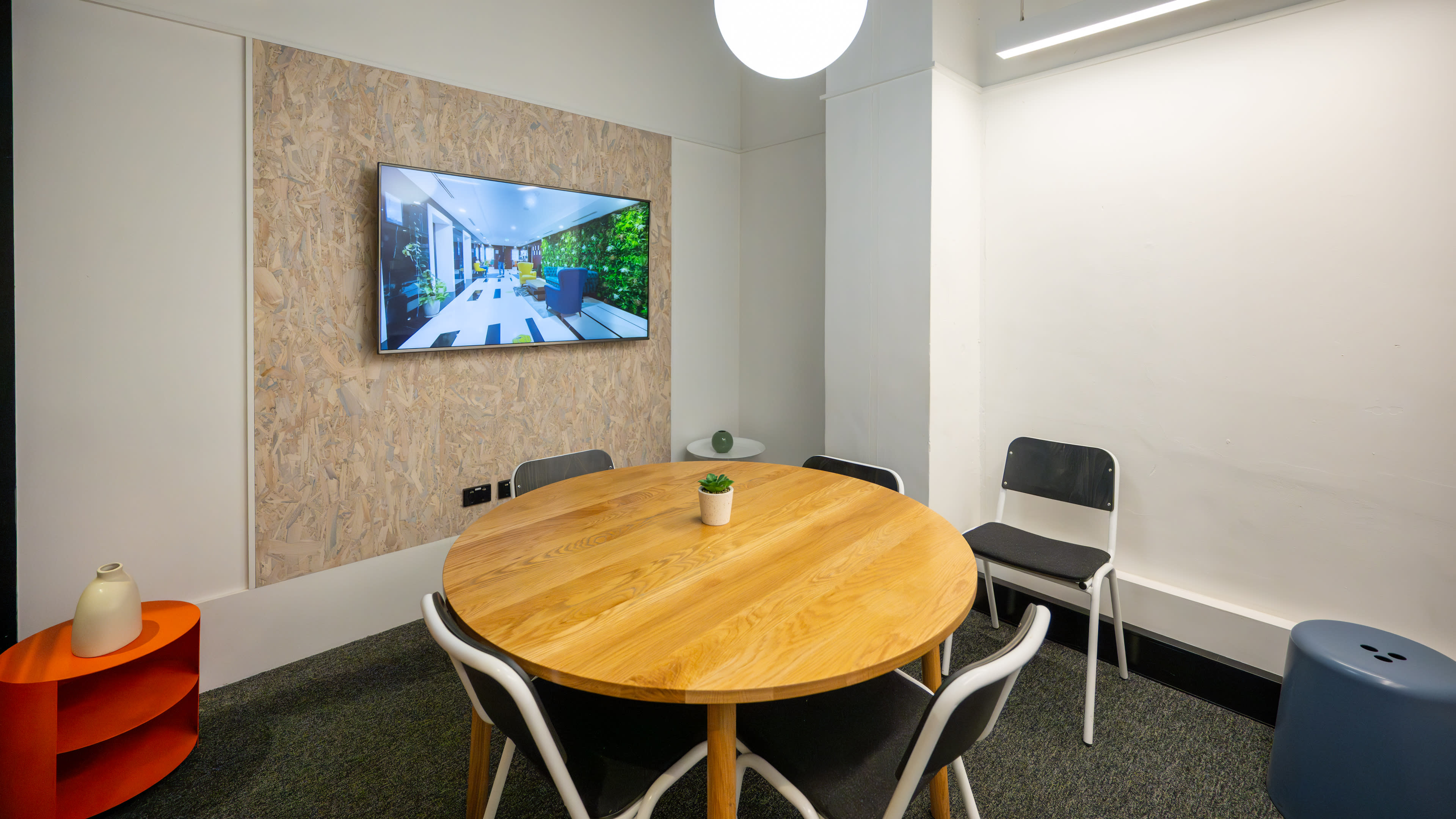 A round wooden table with chairs is set in a modern meeting room featuring a wall-mounted screen displaying a landscaped area.