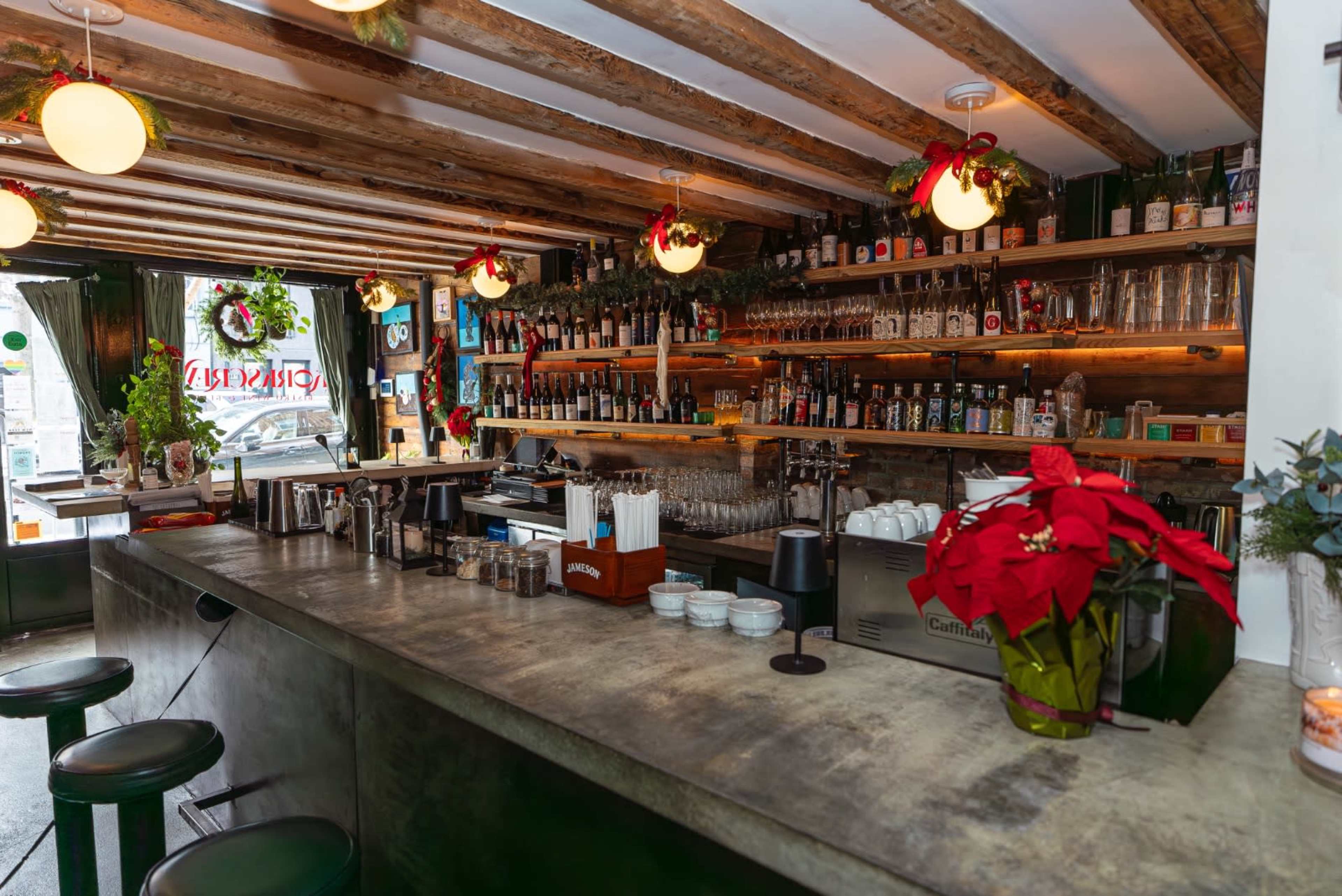 The image shows a cozy bar interior with a concrete counter, wooden beams, and a variety of bottles displayed on shelves behind the bar, decorated with holiday garlands.