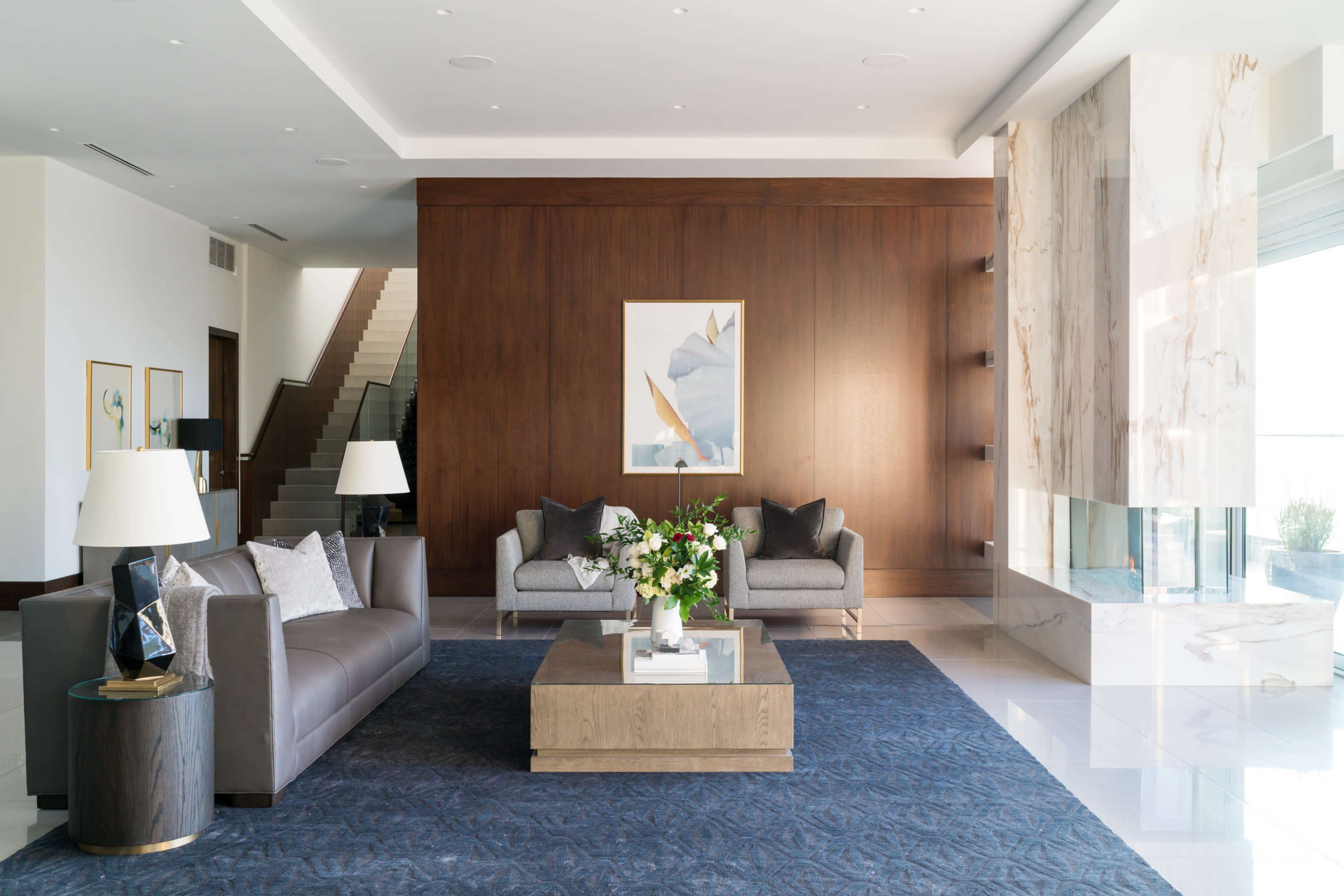 The image shows a modern living room with a gray sofa, a wooden coffee table, and a large artwork on the wooden wall, accompanied by a staircase in the background.