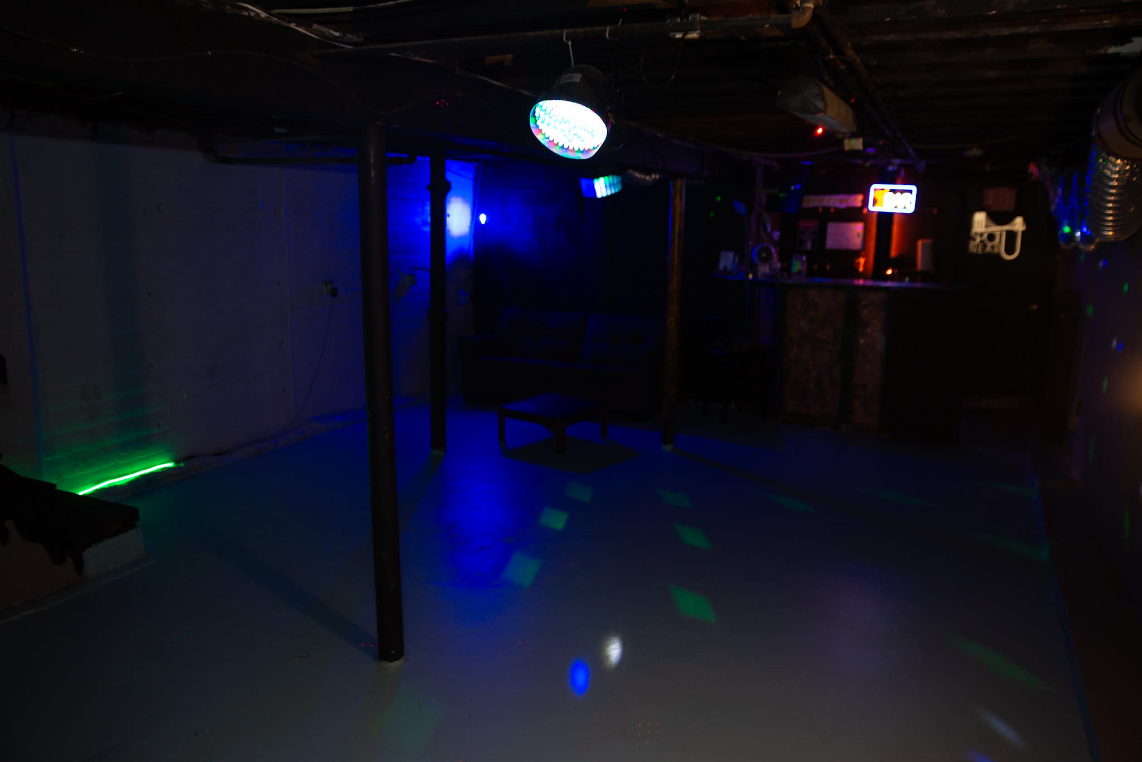 The image shows a dimly lit basement room with colorful LED lights, a bar area in the background, and a couch along one wall.