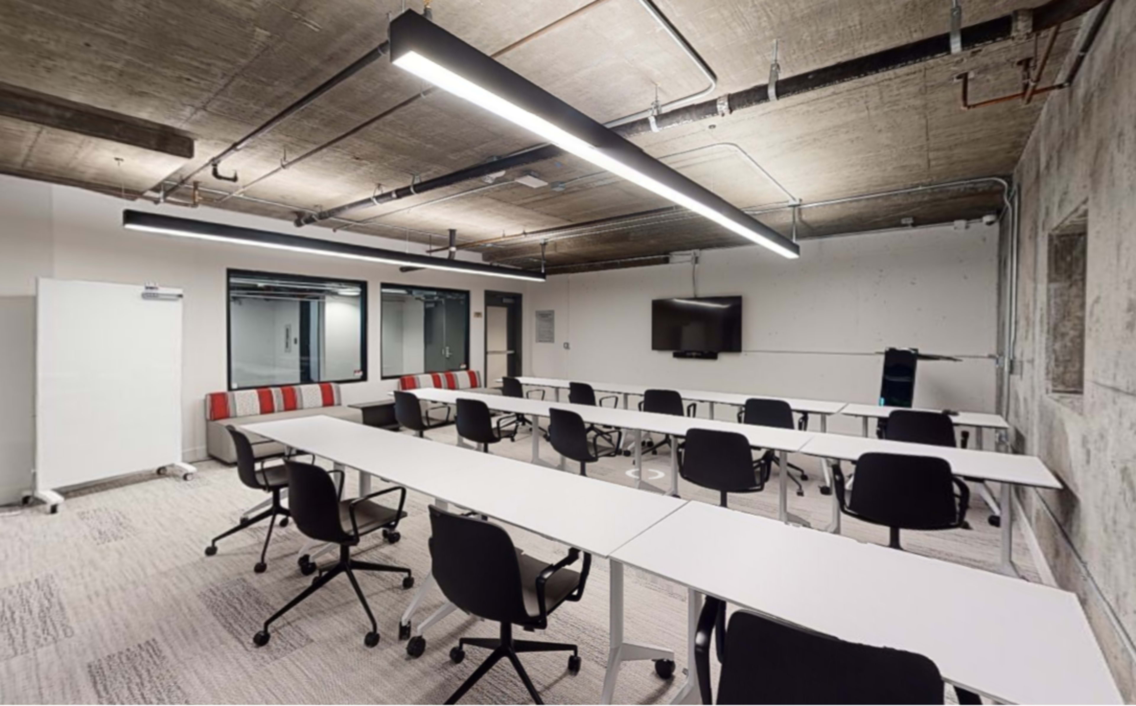 A modern classroom or meeting space with white tables arranged in a U-shape and black chairs, equipped with a television and a glass wall.