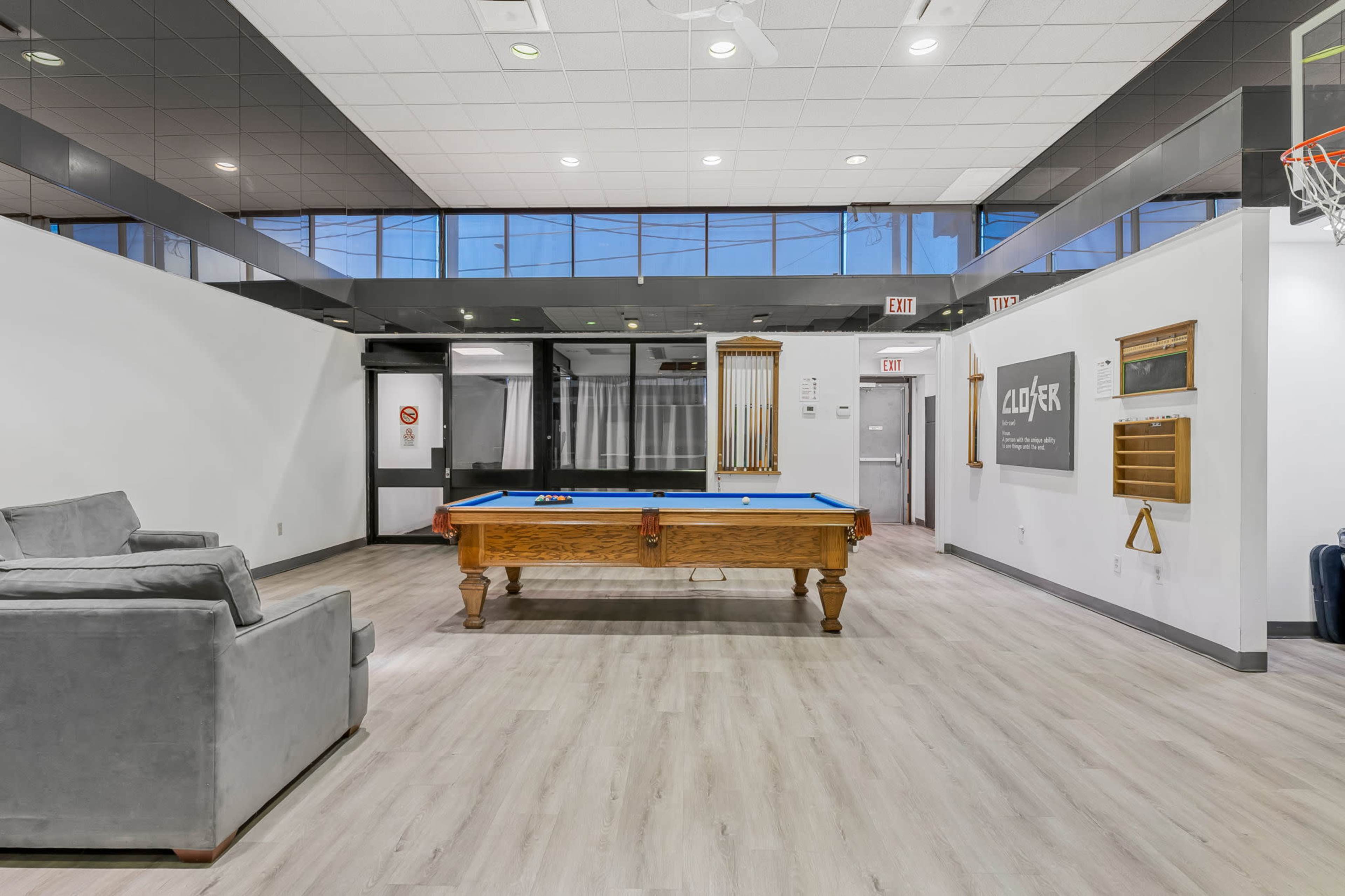 The image shows a recreational room featuring a billiards table, a gray sofa, and large windows allowing natural light to enter.