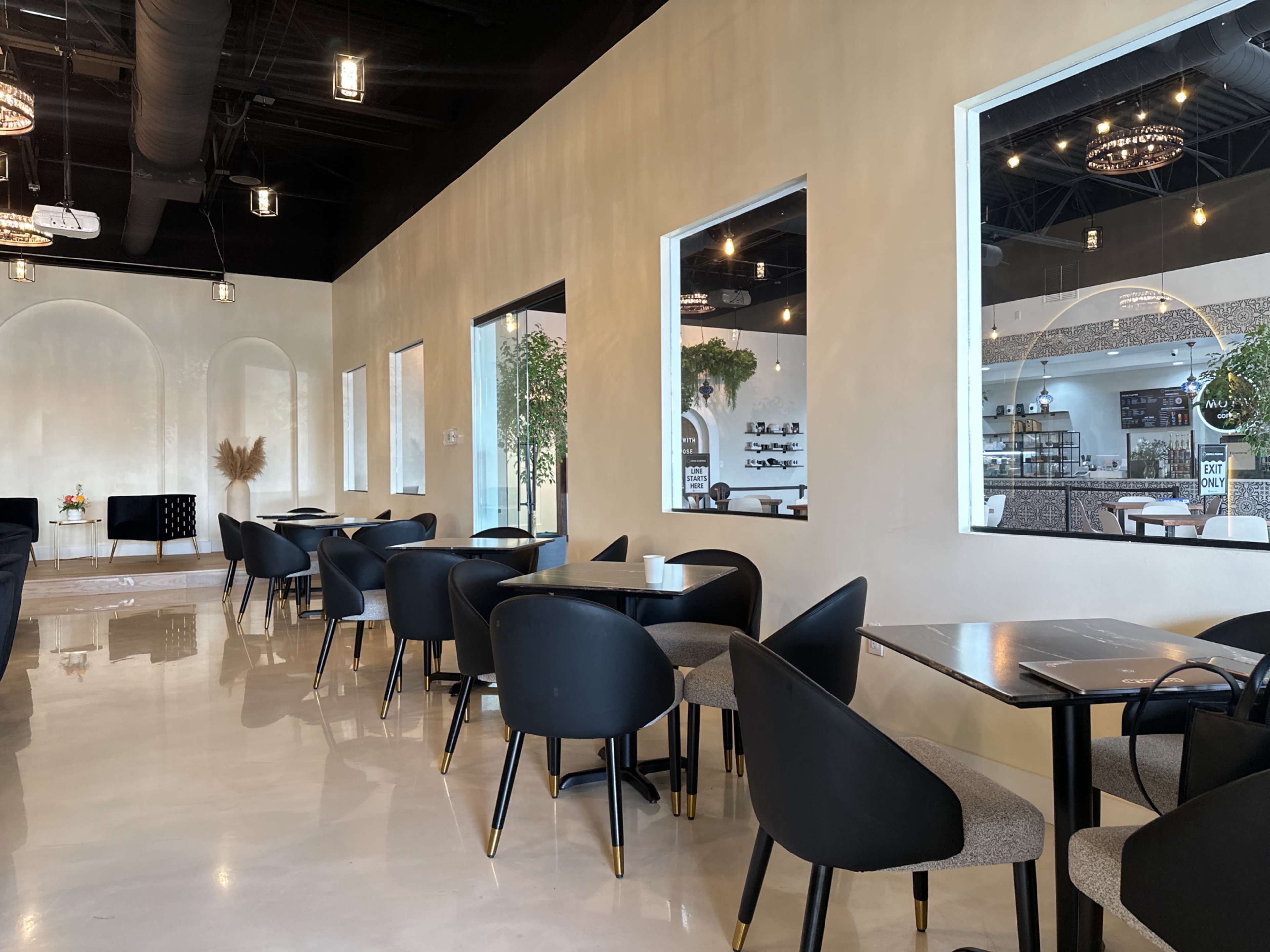 The image shows a modern café interior with black chairs and tables arranged neatly along the walls, featuring large windows that overlook a well-decorated space.