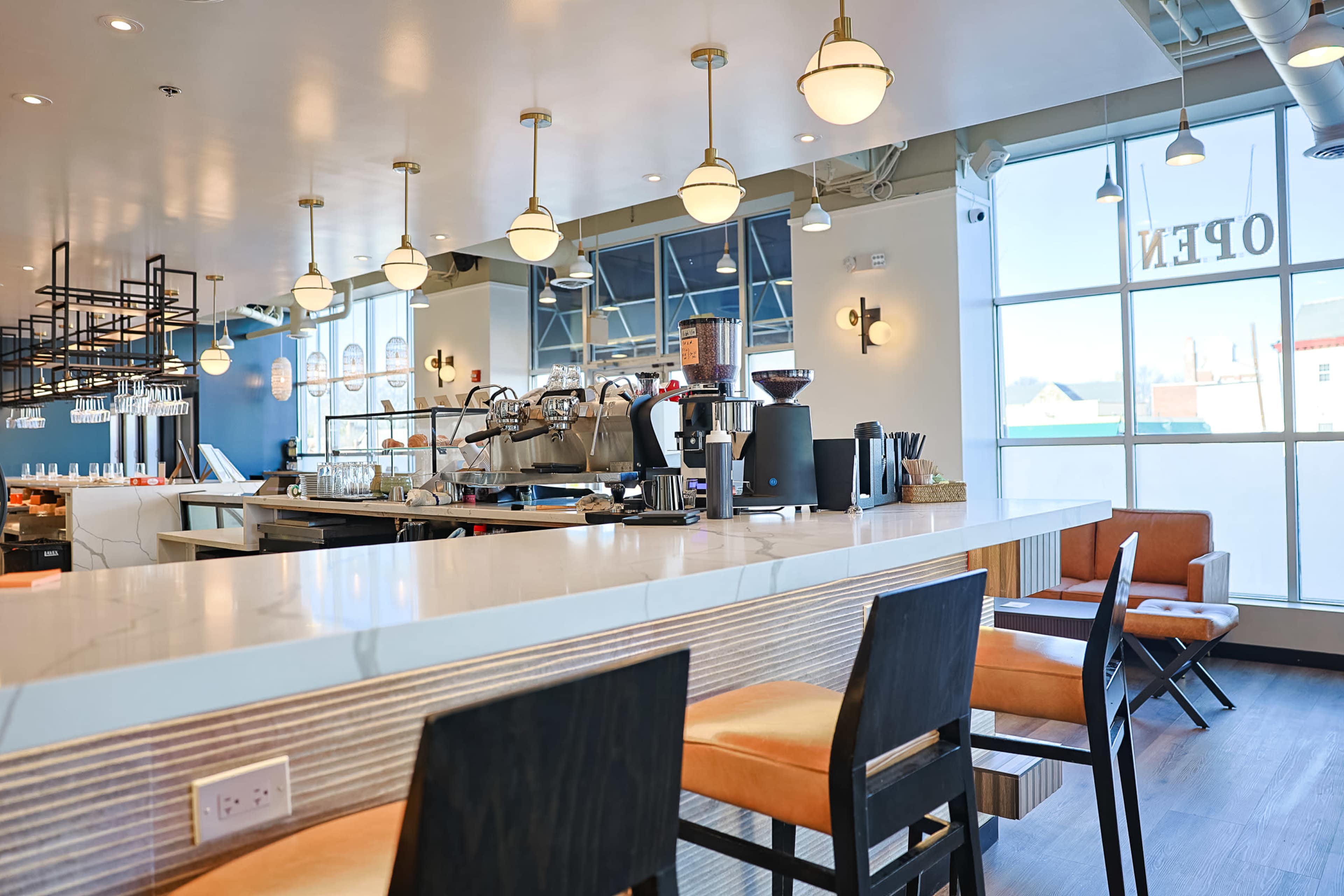 The image shows a modern café interior featuring a long countertop with bar seating, an espresso machine, and bright overhead lighting.