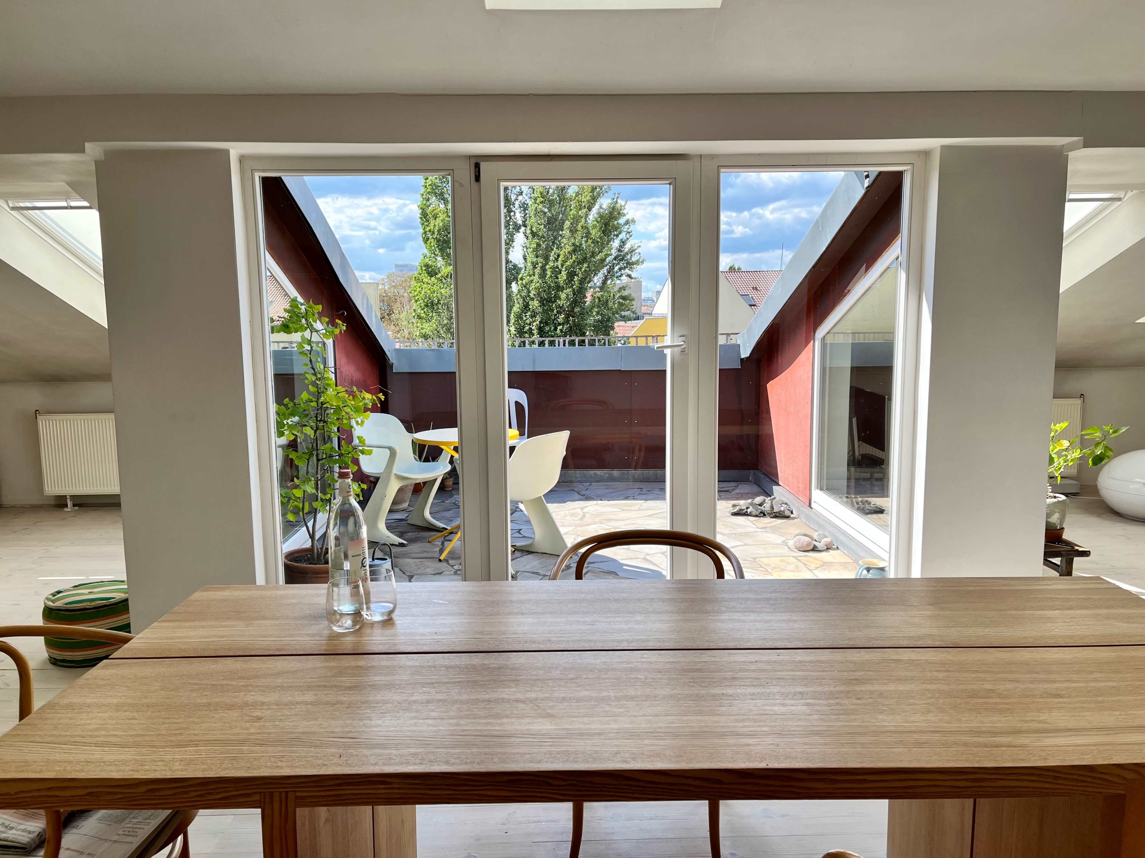 A wooden table is positioned in front of large windows that open to a balcony with outdoor seating and greenery.