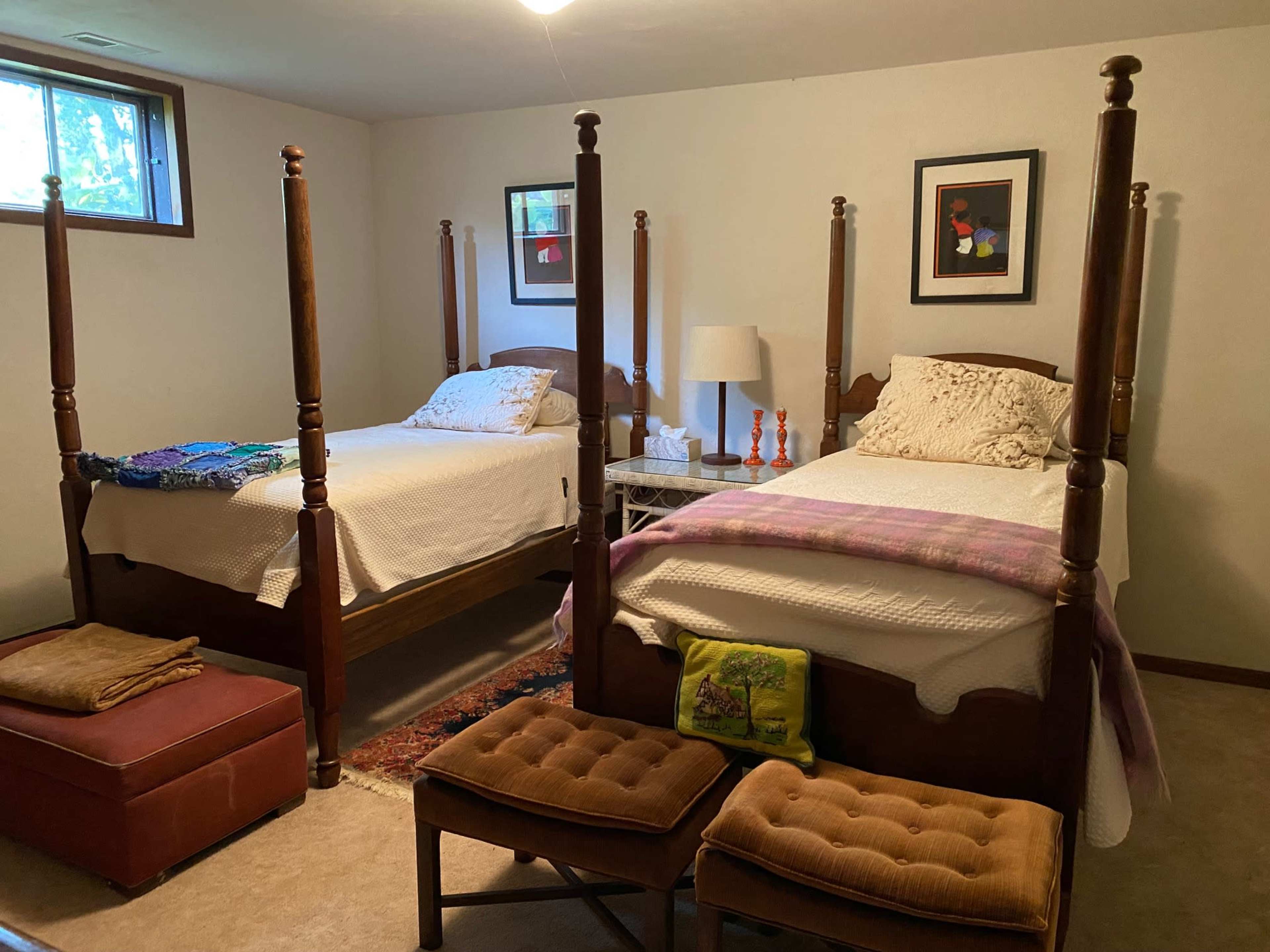 A room with two four-poster beds, a small seating area with ottomans, and a side table with a lamp.