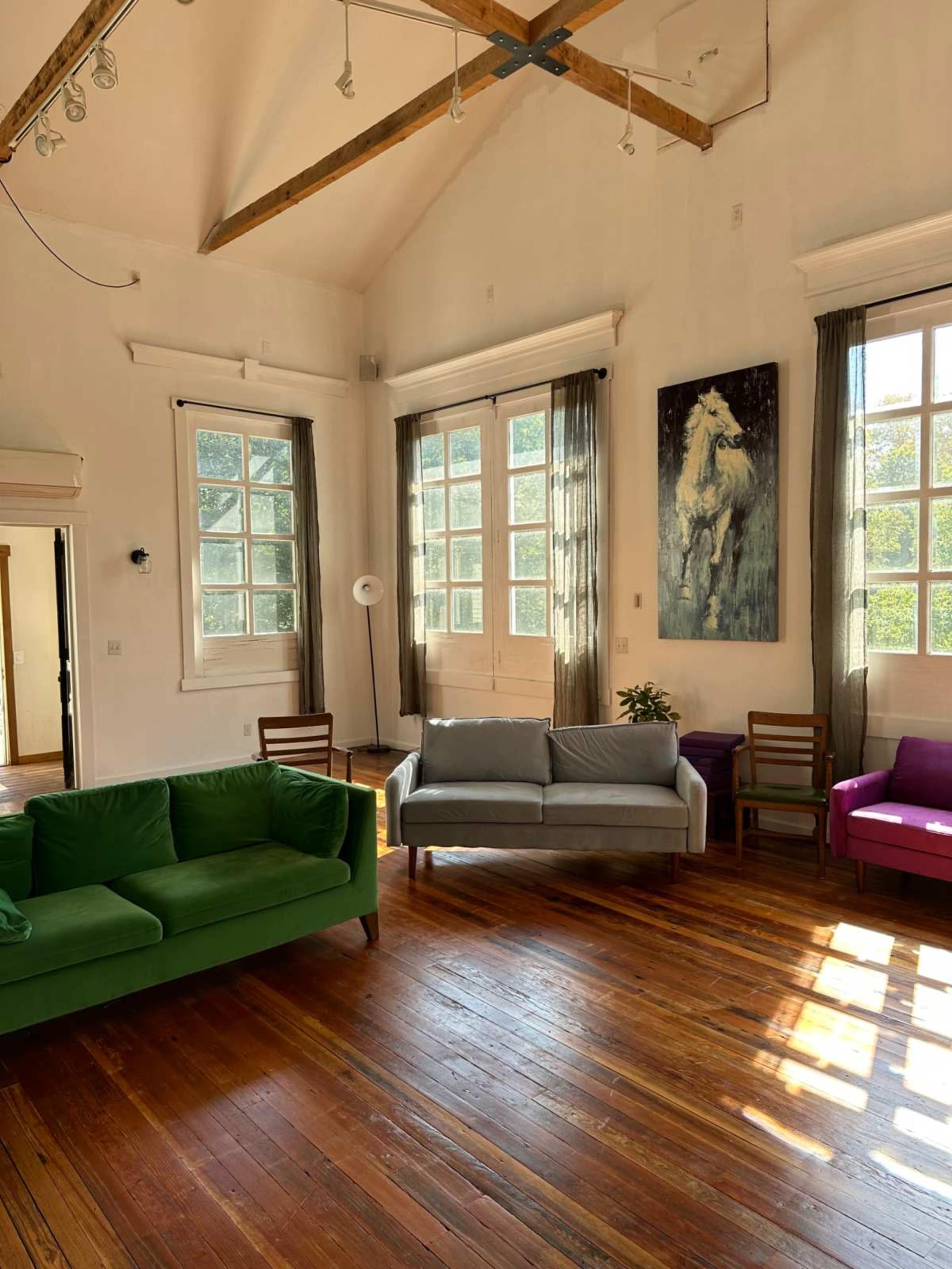 The room features wooden floors, tall windows with natural light, and a mix of green and purple sofas arranged around a central space.
