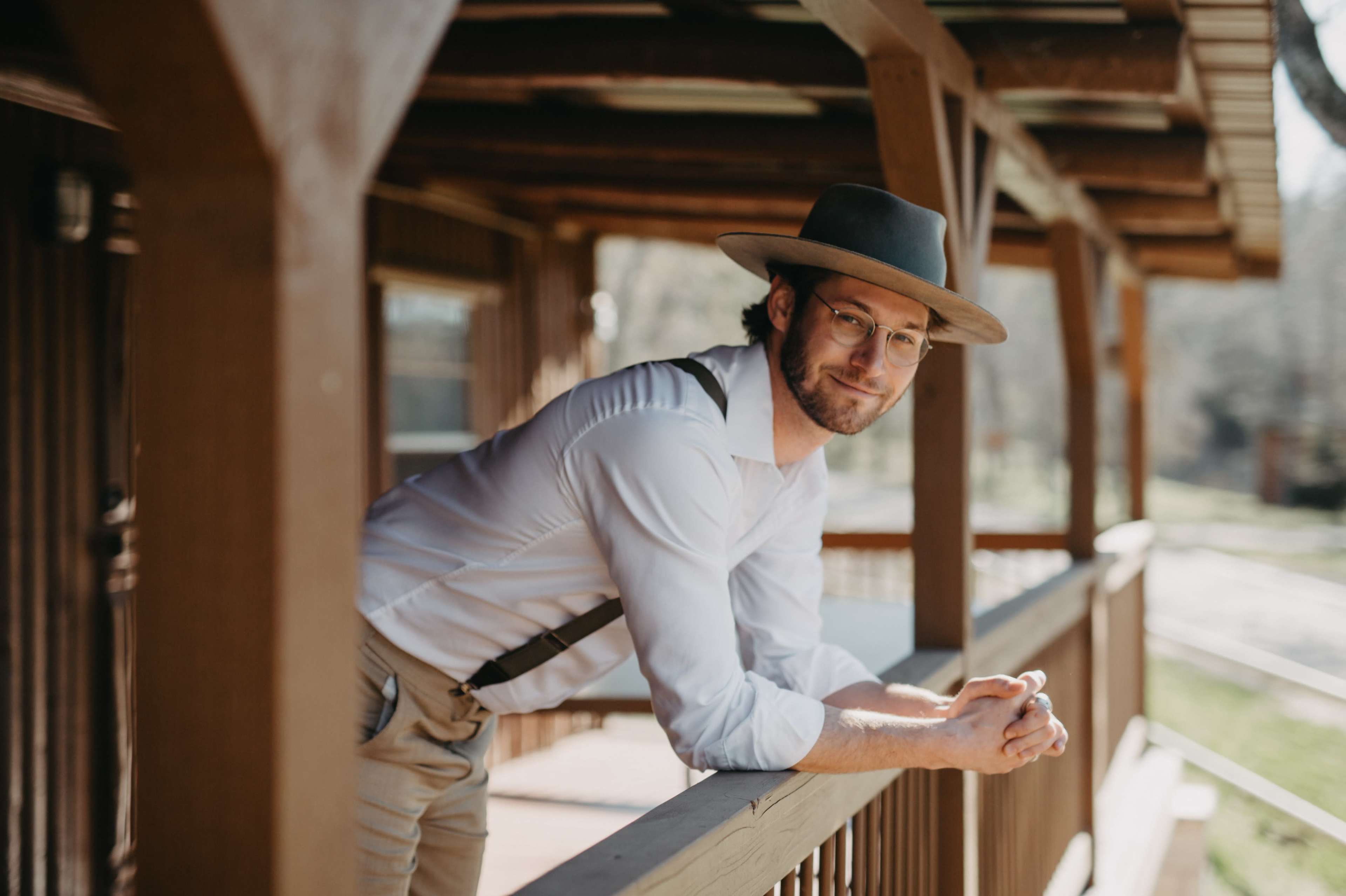 A man in a white shirt and gray hat leans against a wooden railing on a porch.