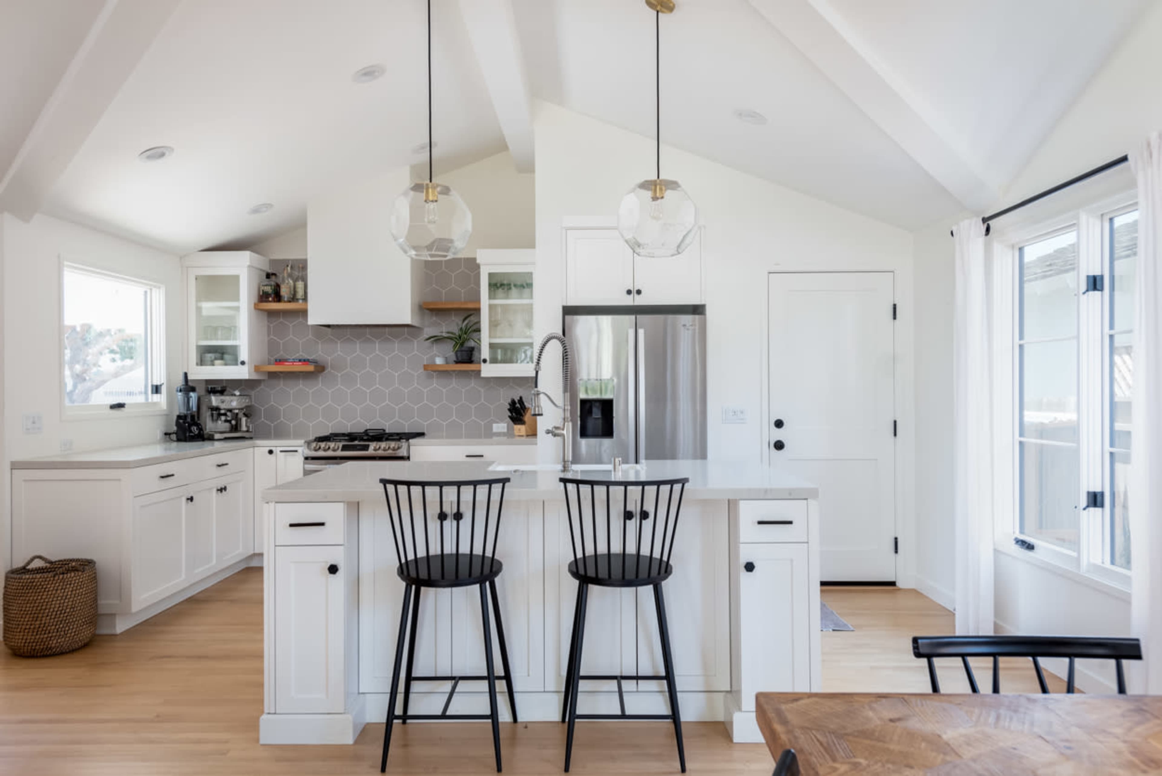 A modern kitchen features white cabinetry, a central island with black bar stools, and large windows allowing natural light to enter.