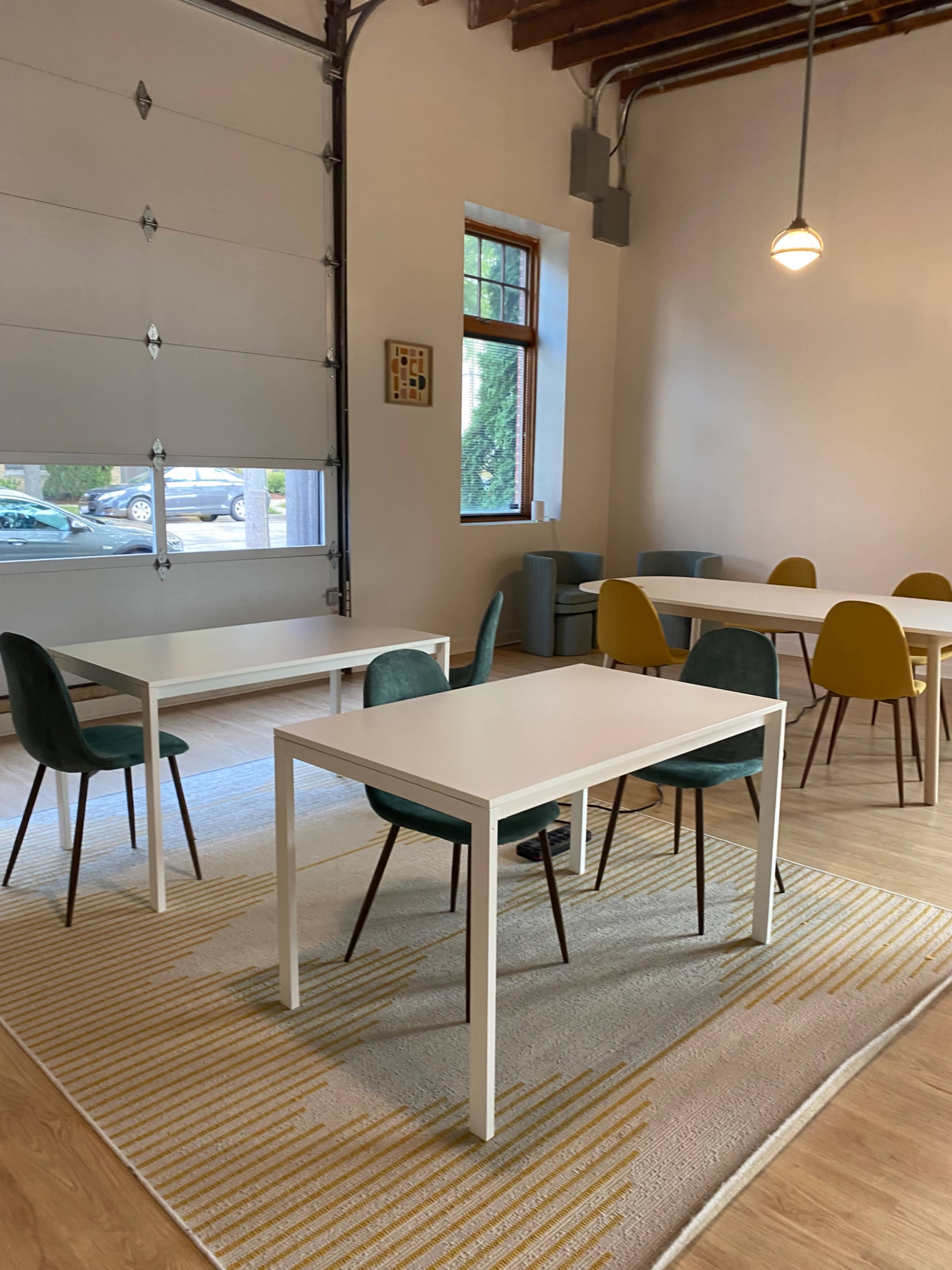 A minimalist room features several tables and chairs arranged on a patterned rug, near a large garage door and a window.