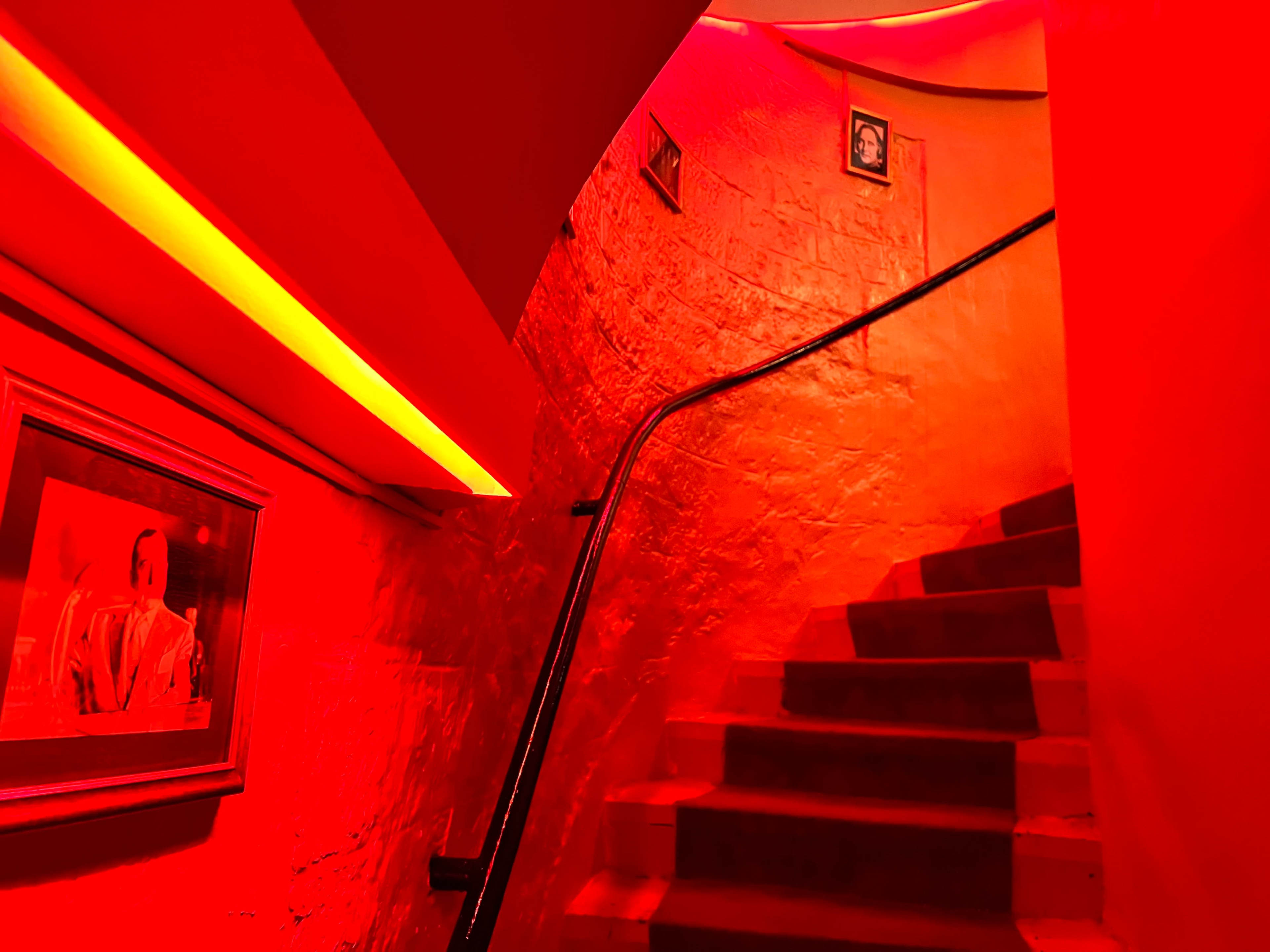 A staircase illuminated by red lighting leads upwards, with framed pictures along the walls.