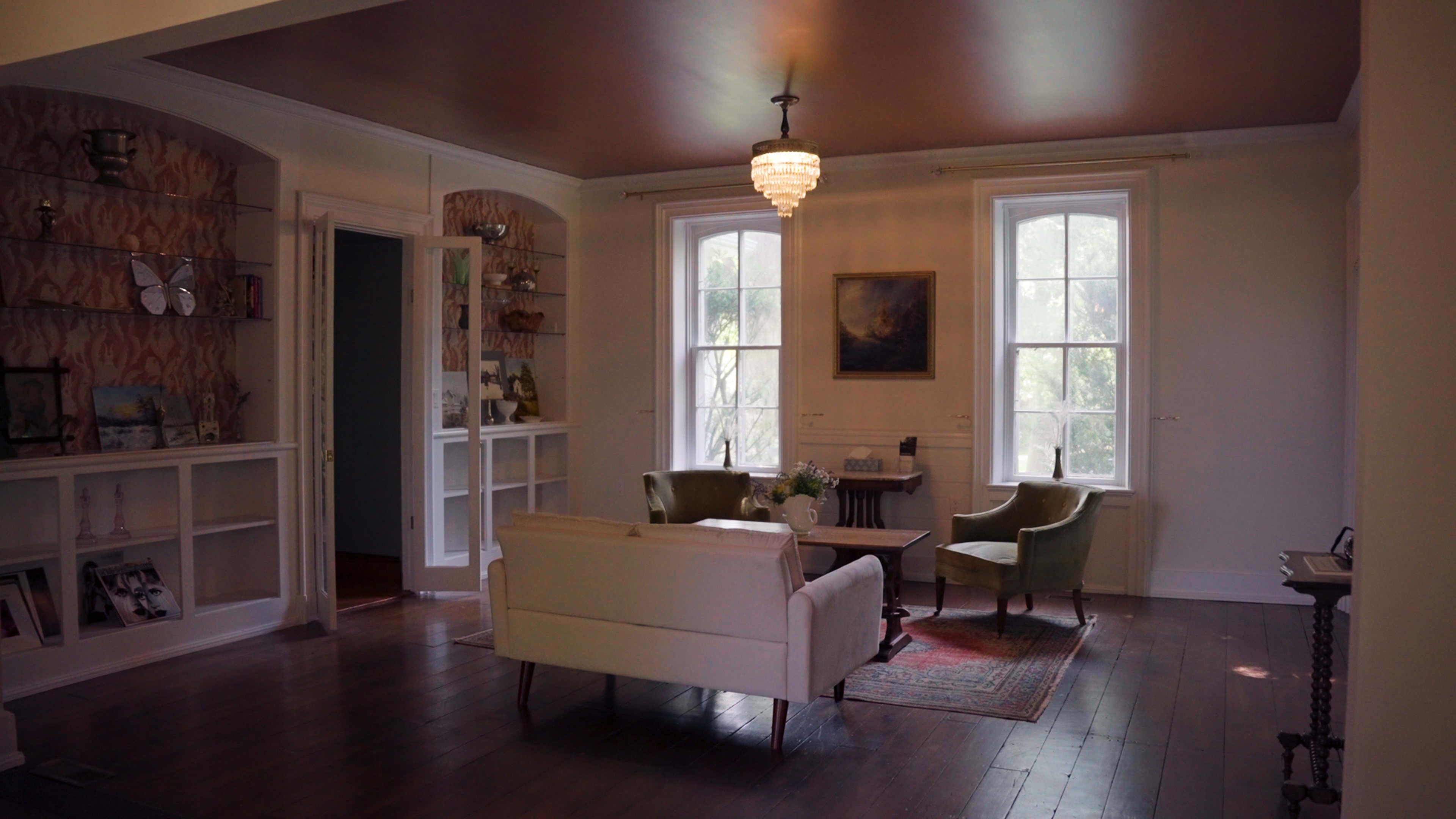The image shows a light-filled room with a white sofa, two green chairs, and large windows alongside decorative shelving.