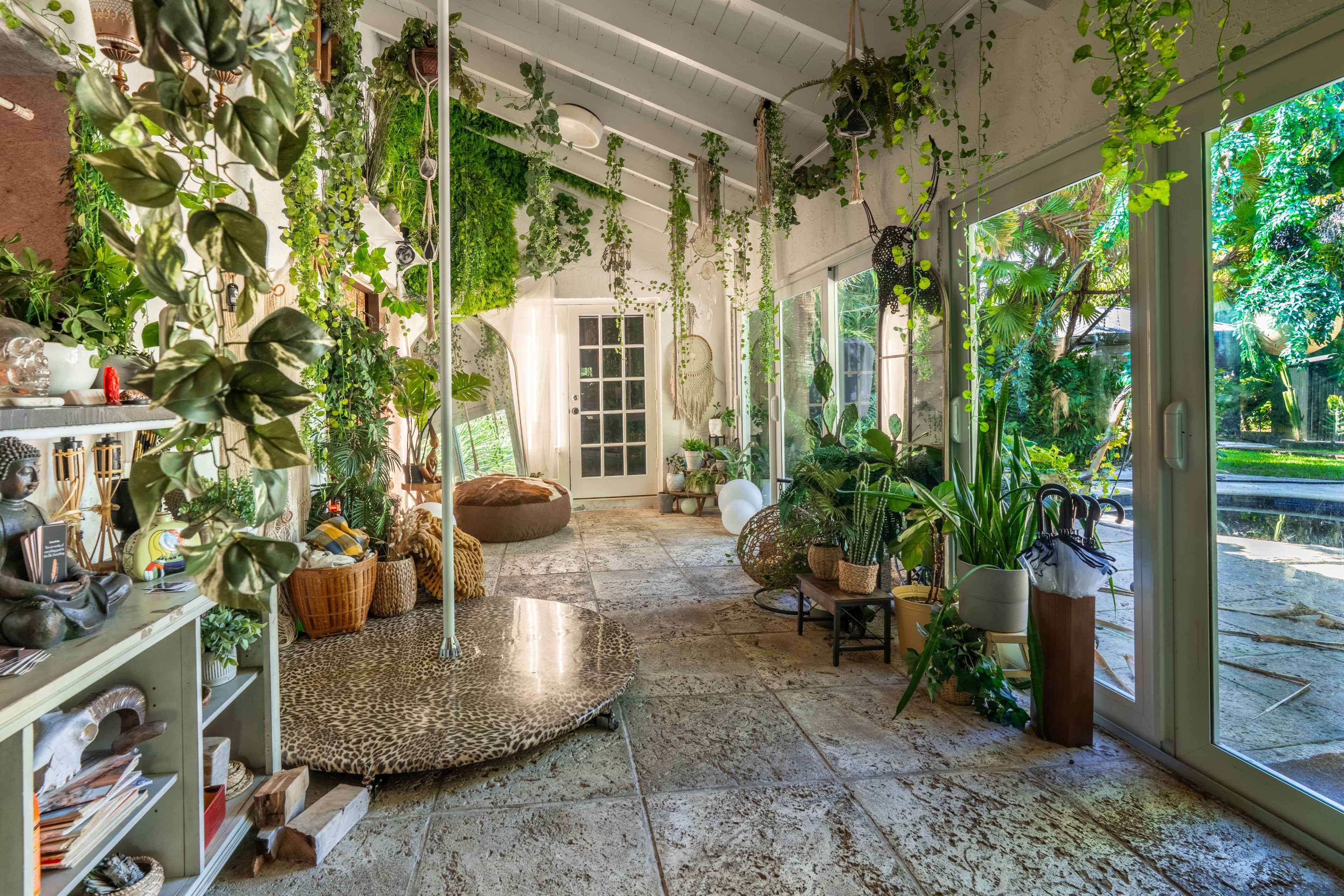 The image shows a sunlit indoor space filled with various plants, a comfortable seating area, and large glass doors opening to an outdoor garden.