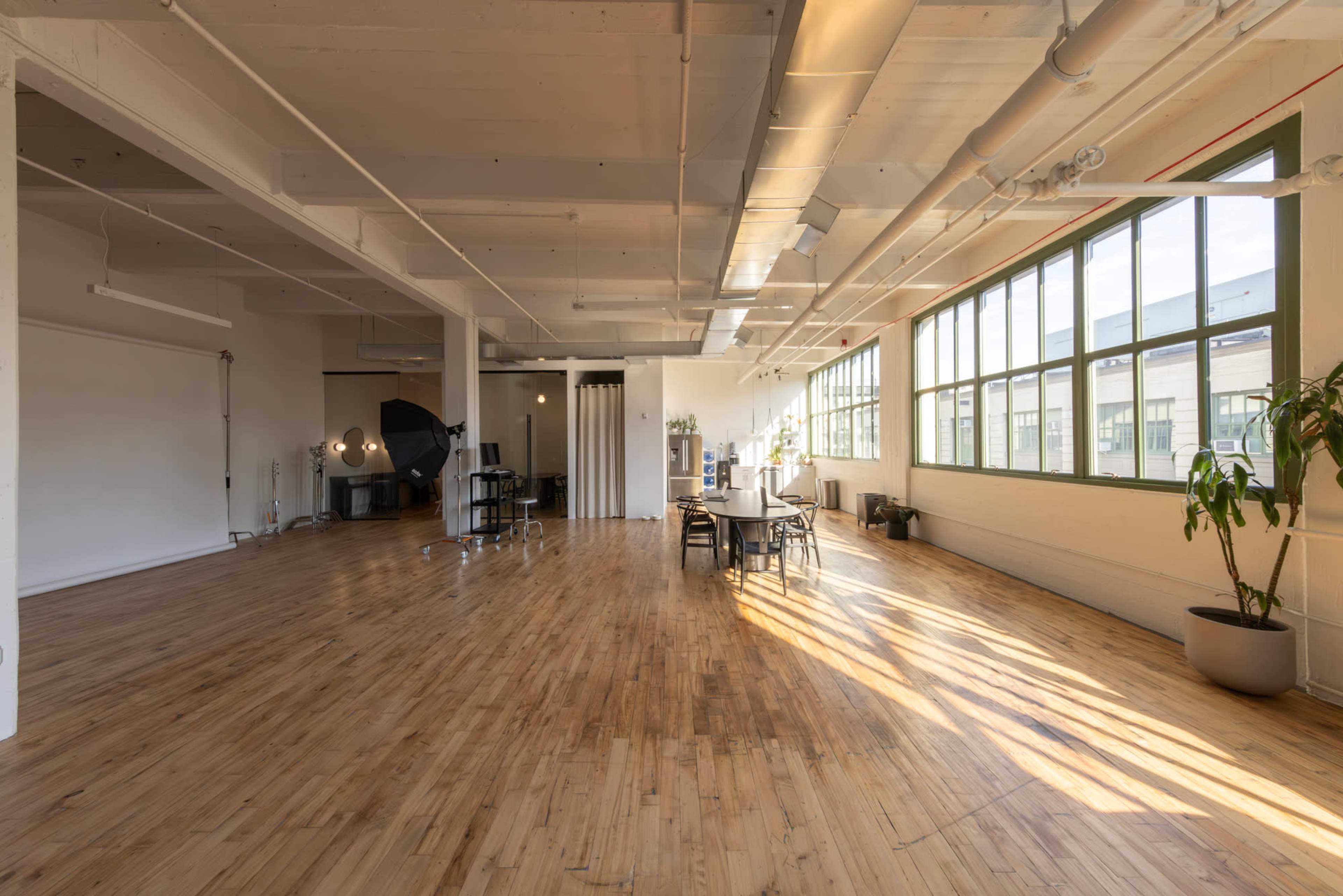 The image shows a spacious, sunlit studio with wooden flooring, large windows, and minimal furniture, including chairs and a table.