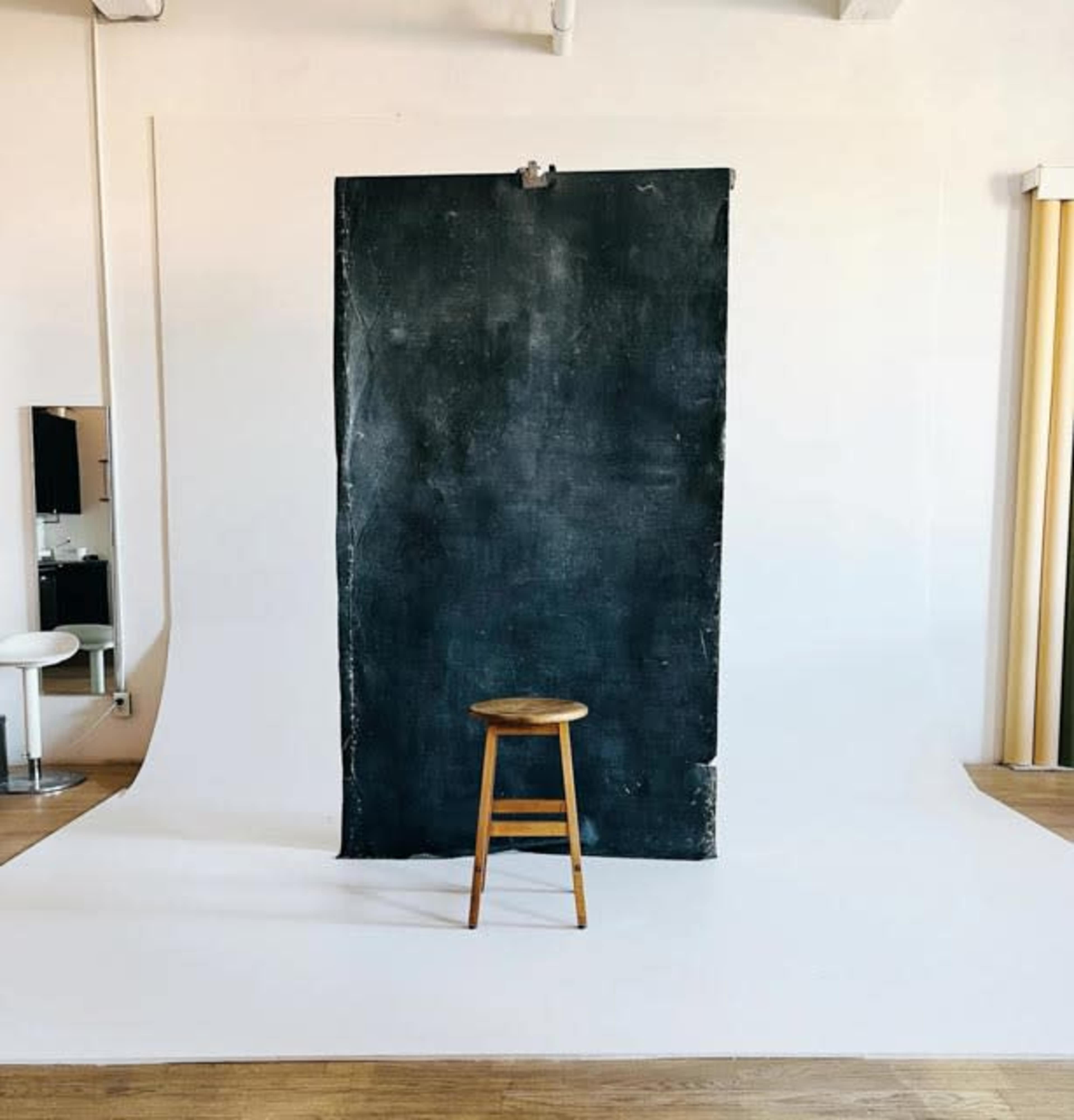 The image shows a minimalist photography studio featuring a black backdrop, a wooden stool, and a white floor.