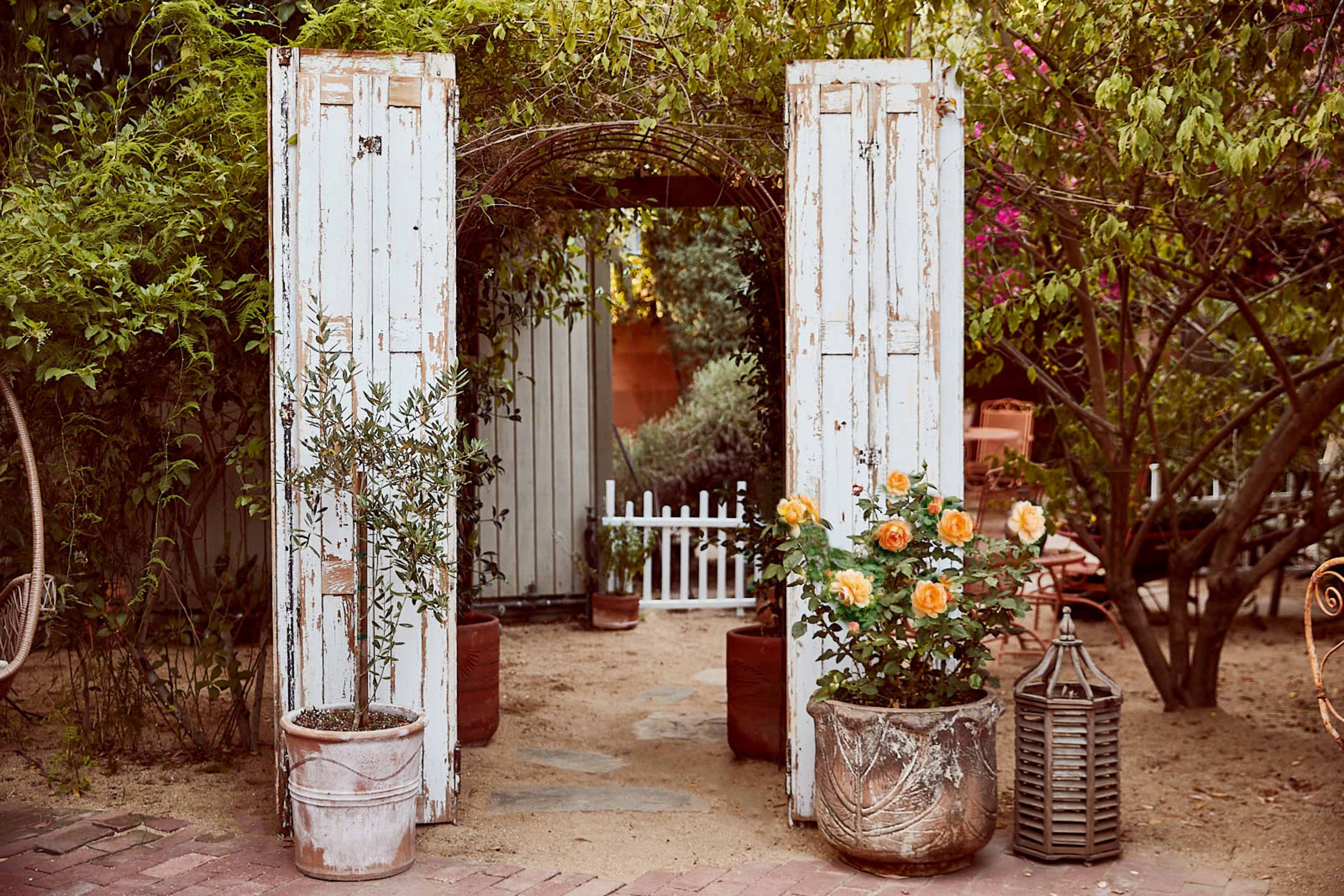 An outdoor garden entrance framed by two weathered white doors, with potted plants on either side and a small white picket fence visible in the background.