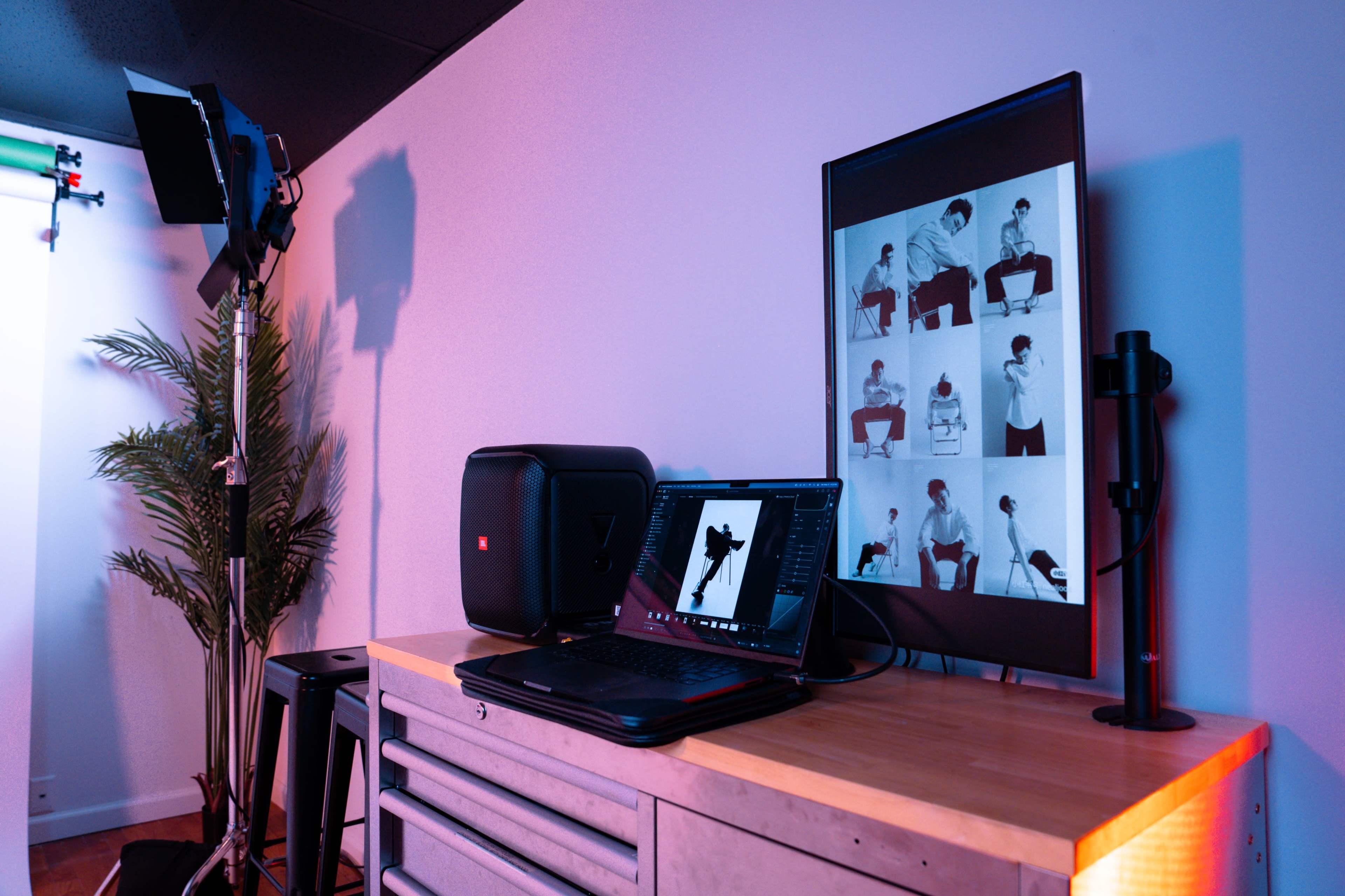 The image shows a work setup in a studio with a computer monitor displaying black-and-white images, a laptop on a wooden table, and studio lighting equipment nearby.