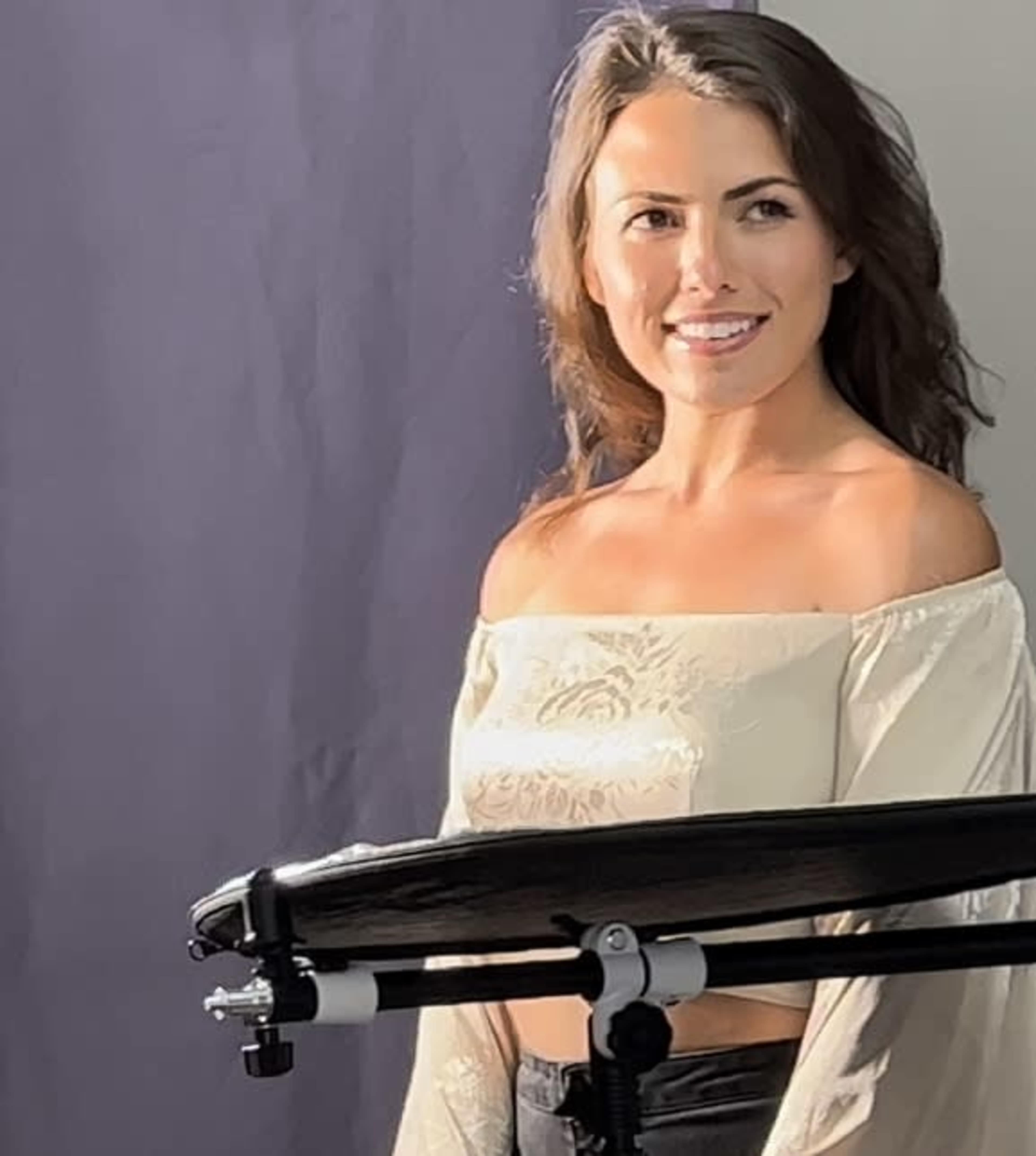 A woman with long hair smiles while standing next to a microphone stand against a backdrop.