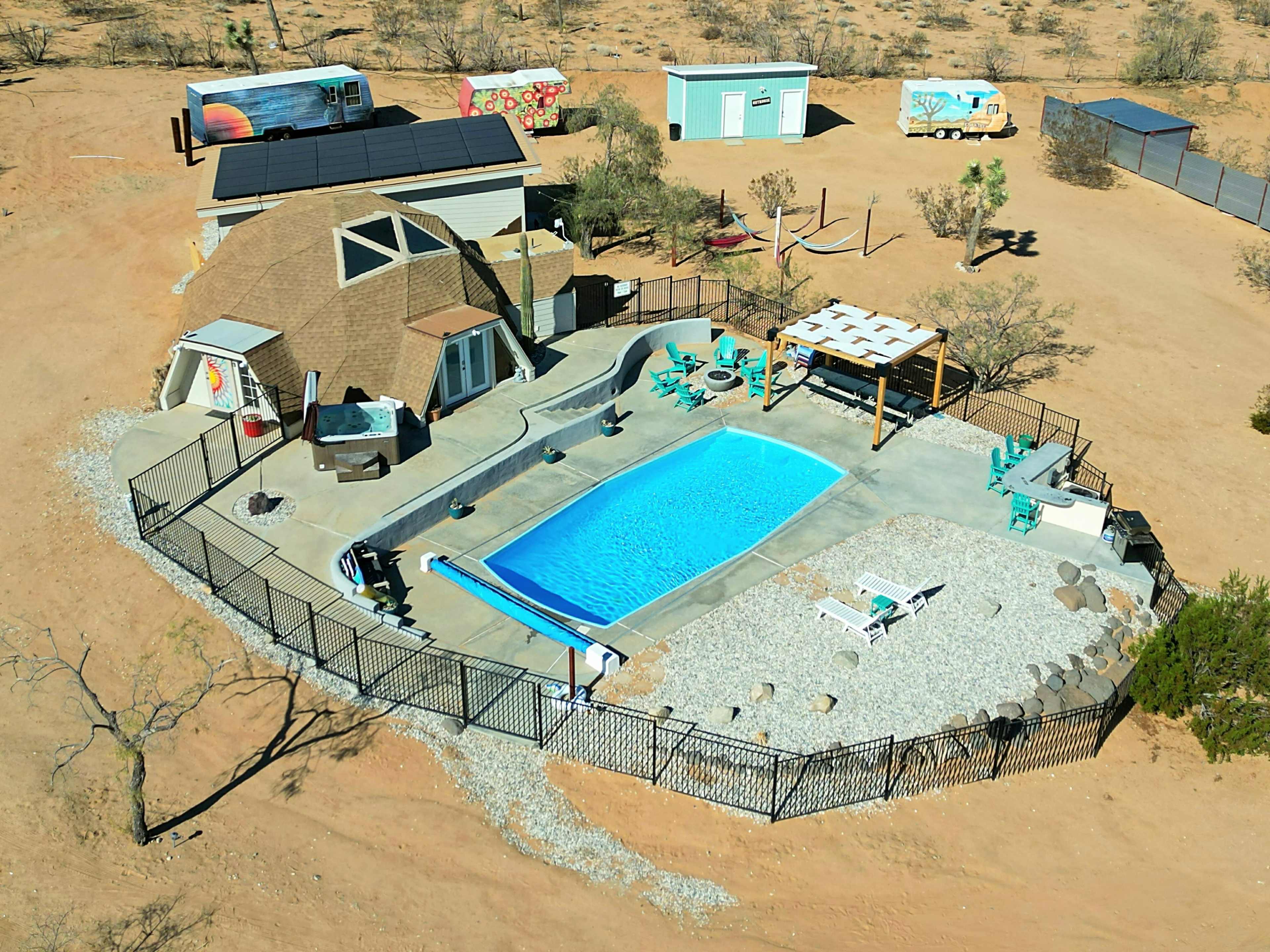 A desert property featuring a dome-shaped house, a swimming pool surrounded by a fence, a shaded seating area, and various outdoor structures.