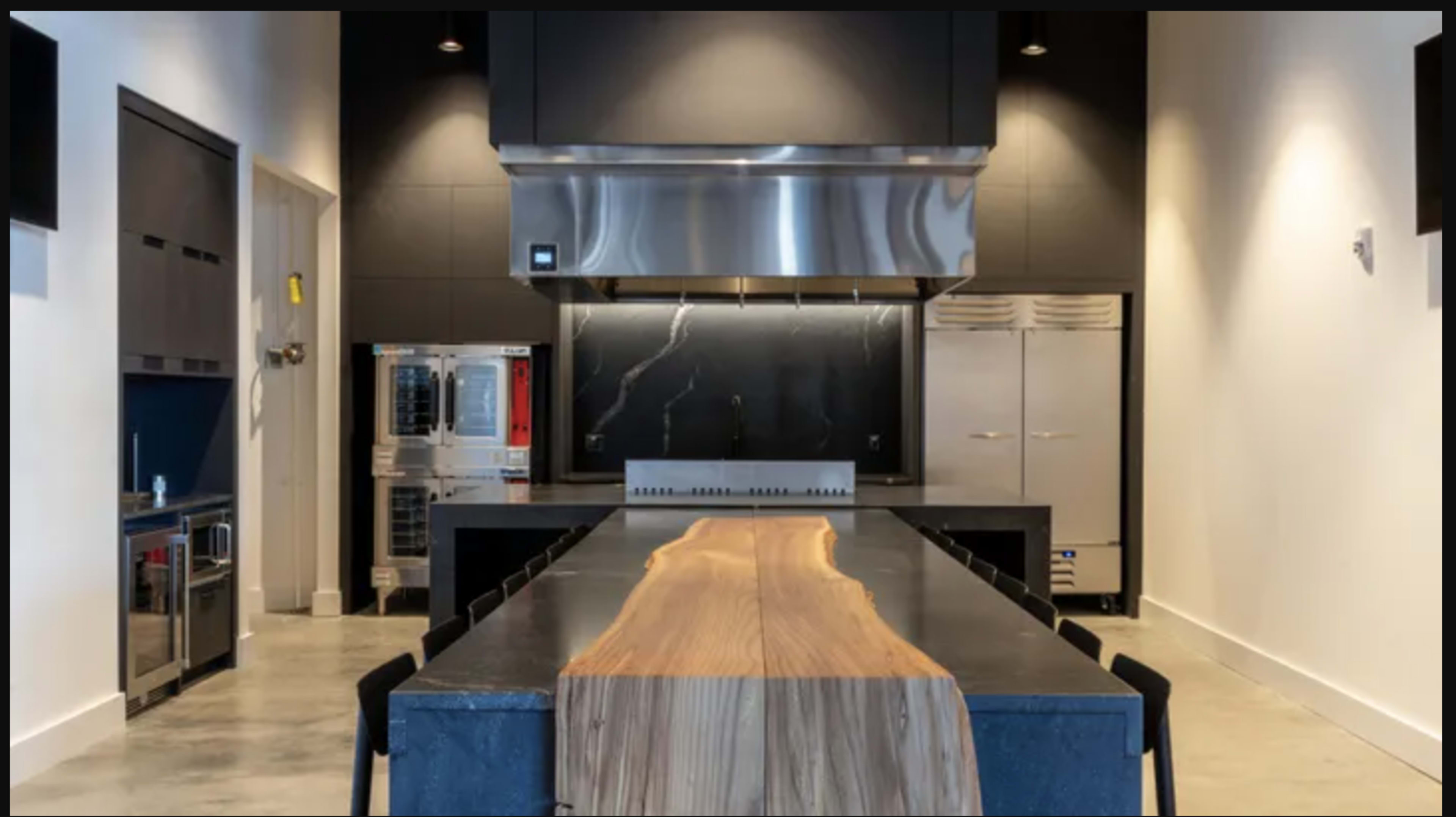 The image shows a modern kitchen featuring a central wooden table with a sleek countertop, stainless steel appliances, and a prominent vent hood above the cooking area.