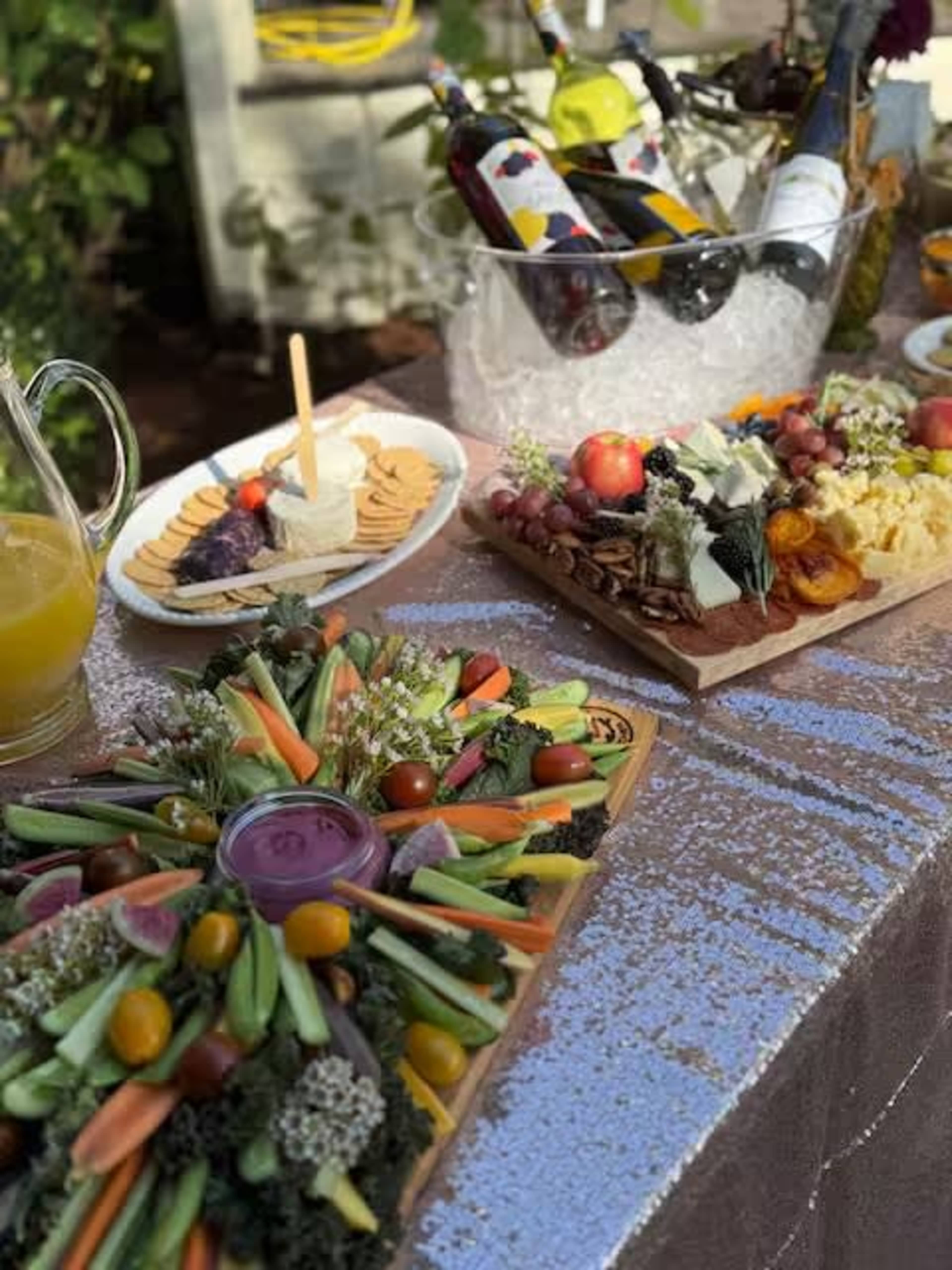 The image features a table set with an array of colorful food platters, including a vegetable platter, cheeses, fruits, and beverages on ice.