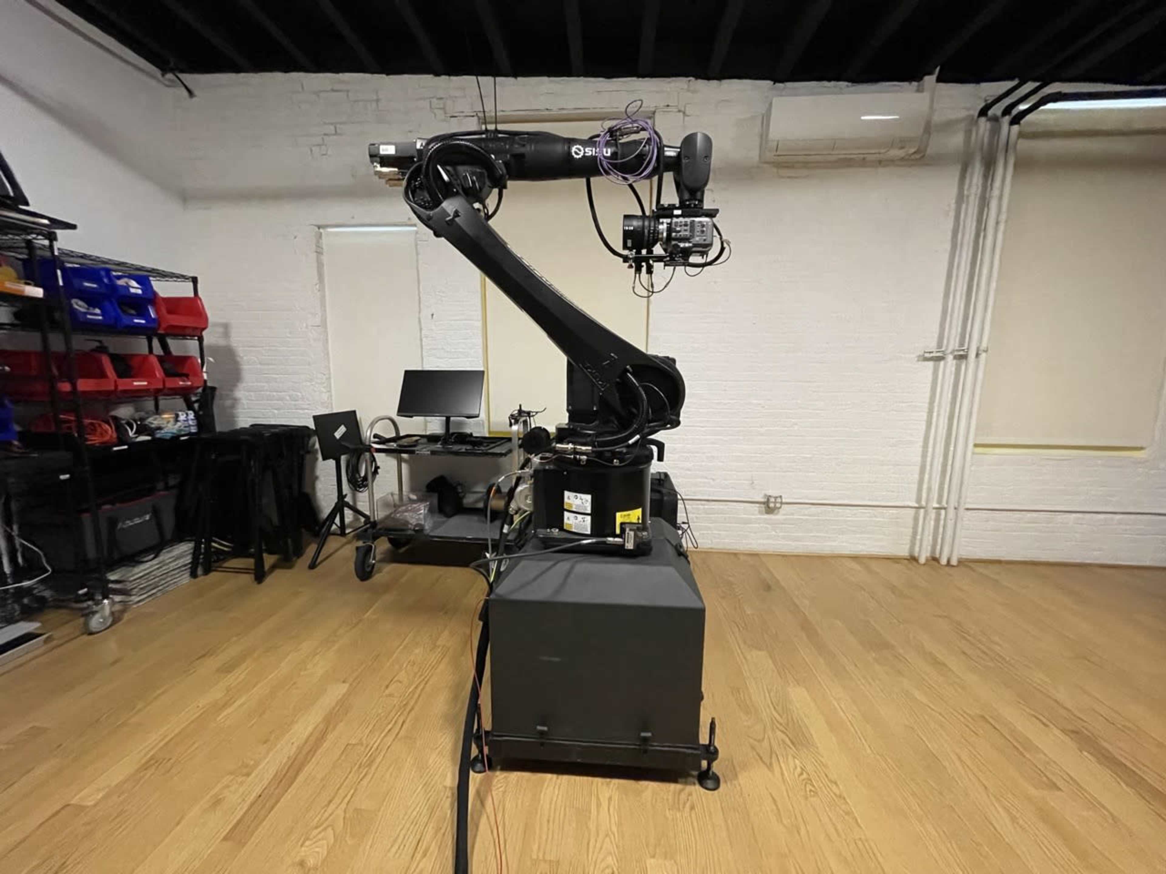 A robotic arm stands on a black base in a spacious room with wooden flooring and various tools and equipment in the background.