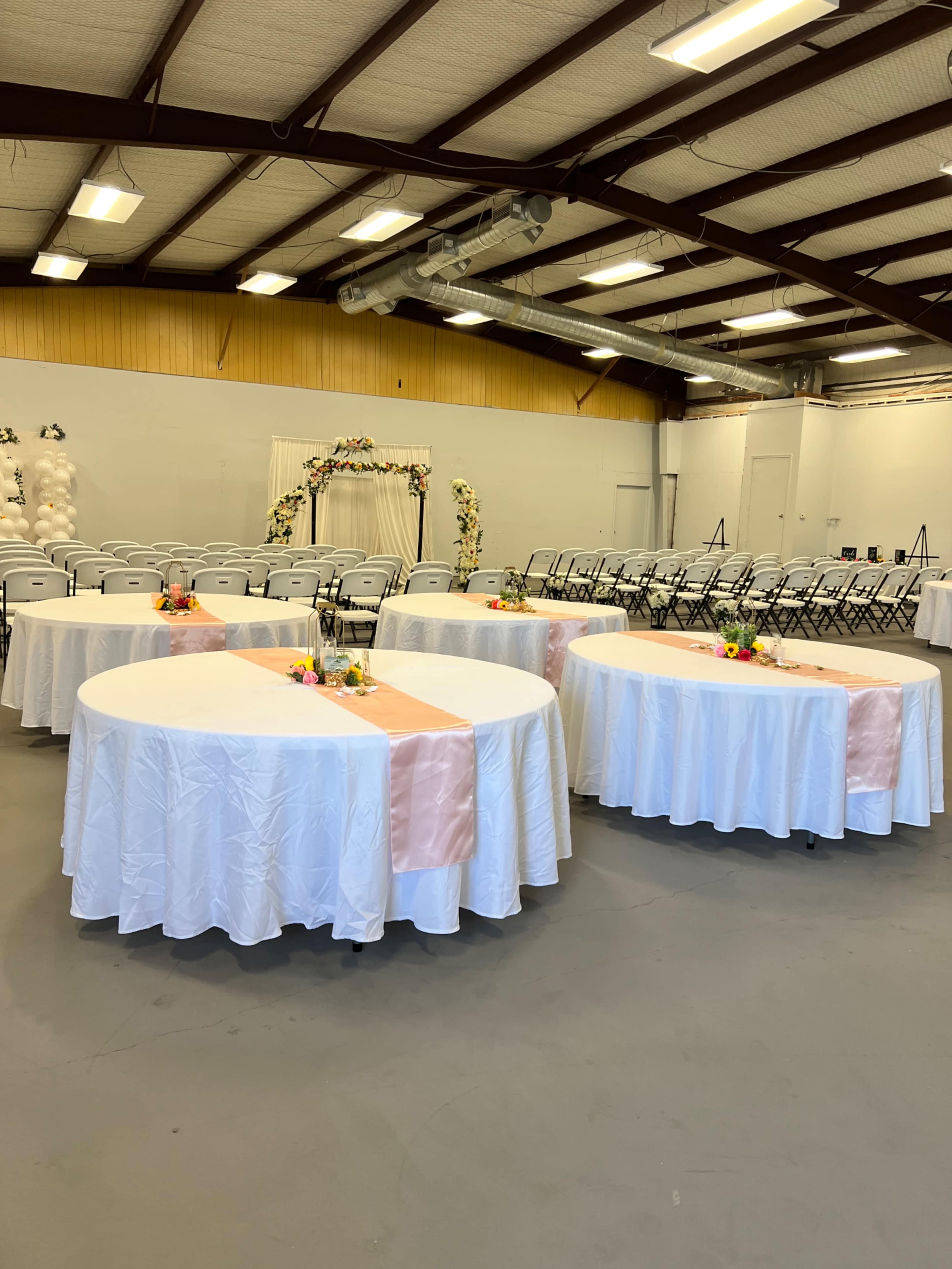 The image shows a spacious indoor event venue with circular tables covered in white tablecloths, decorated with floral centerpieces, and arranged around an area with chairs and a decorative arch in the background.