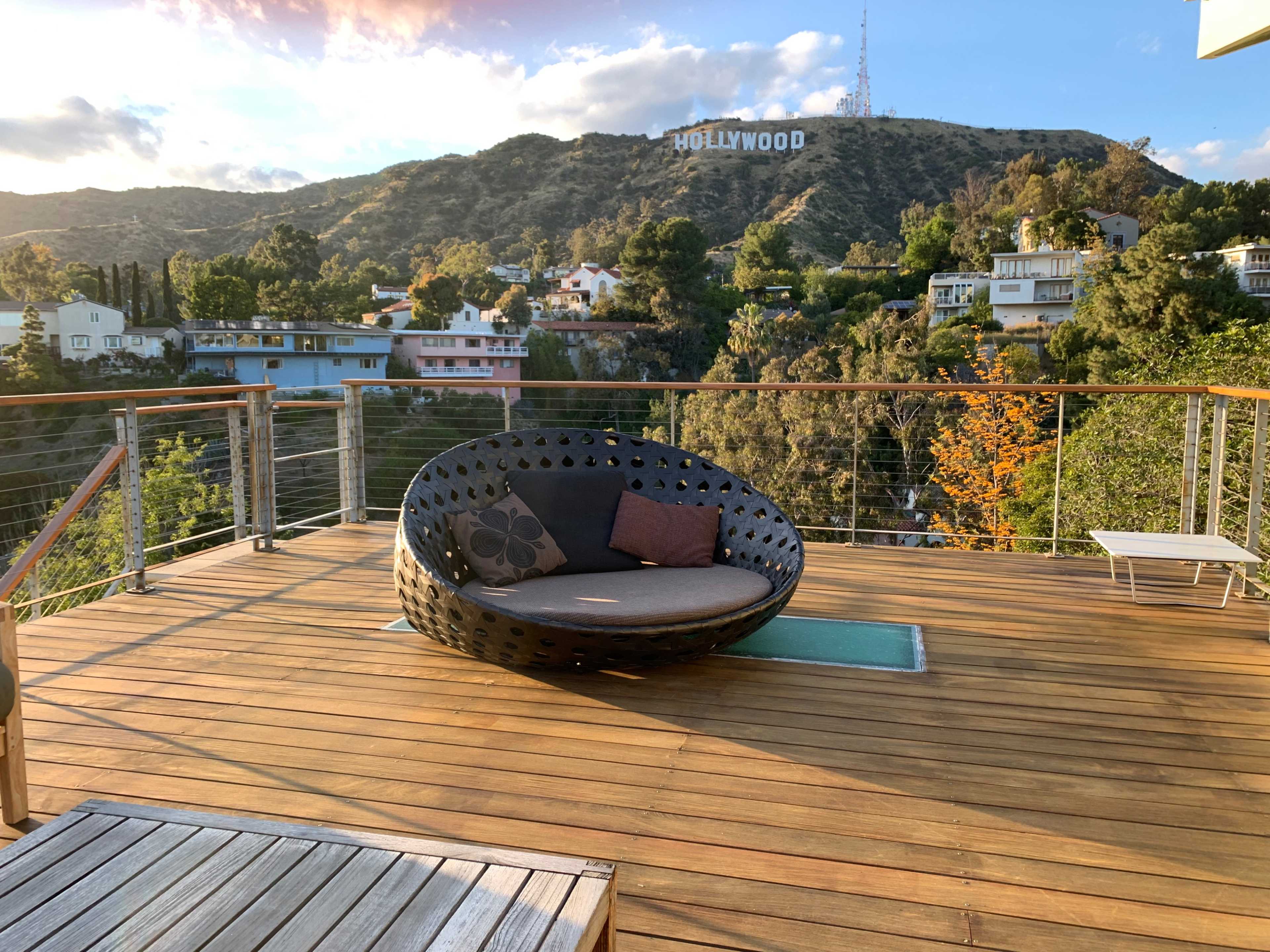 A round, black wicker chair with cushions is situated on a wooden deck overlooking the Hollywood sign on a hillside.