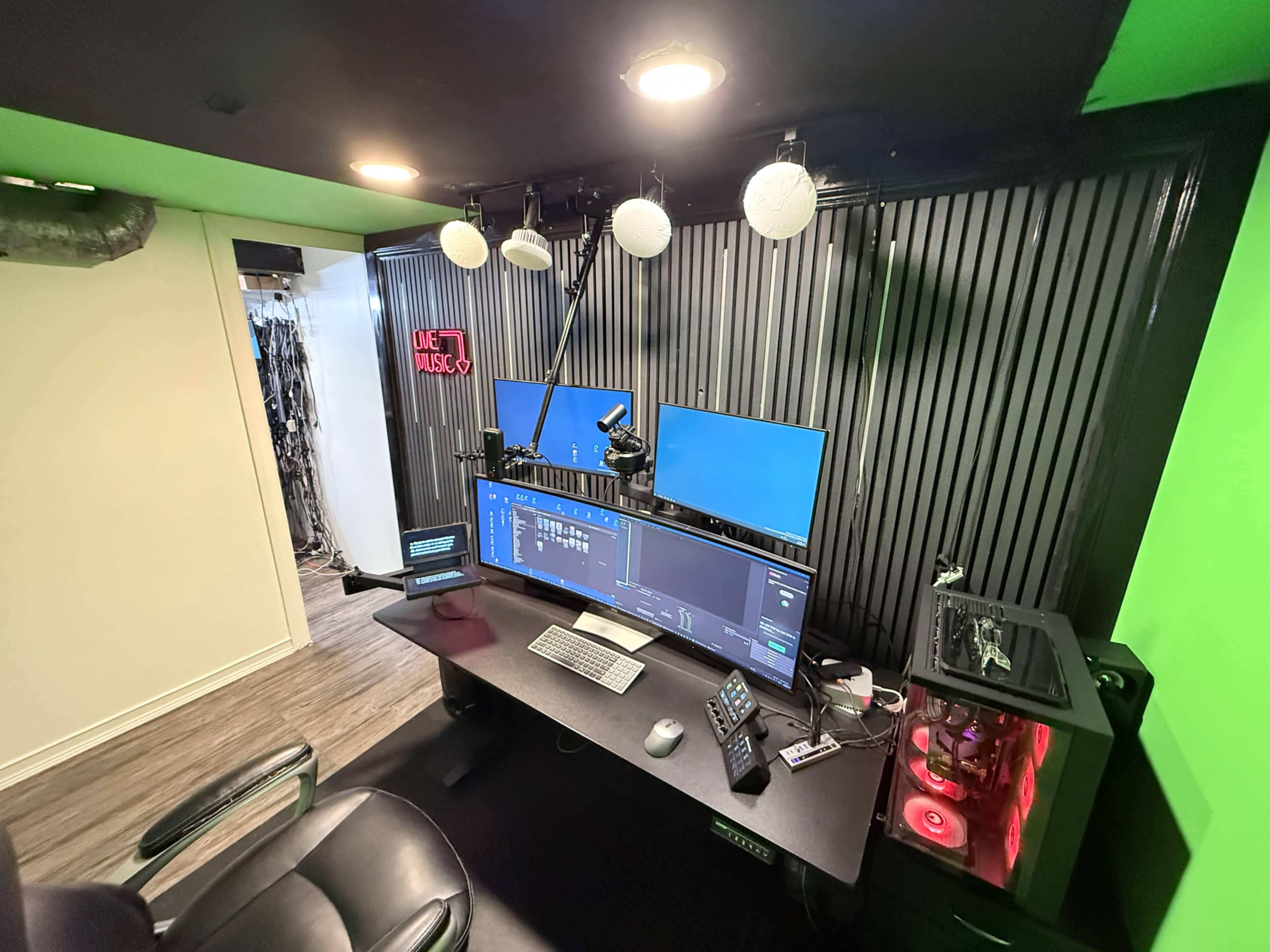 The image shows a modern home office setup with dual monitors, a keyboard, a microphone, and colorful lighting against a green wall.
