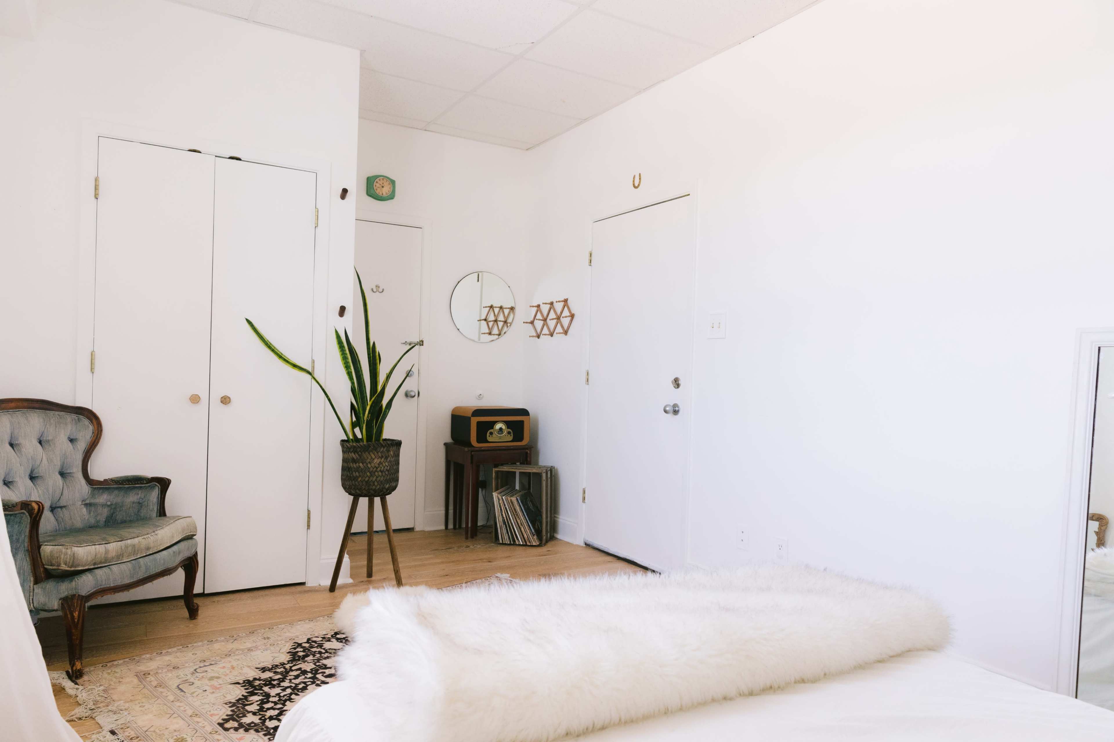 The image shows a bright bedroom with a white wall, a bed adorned with a fluffy throw, a retro record player, a plant in a pot, and a vintage chair.
