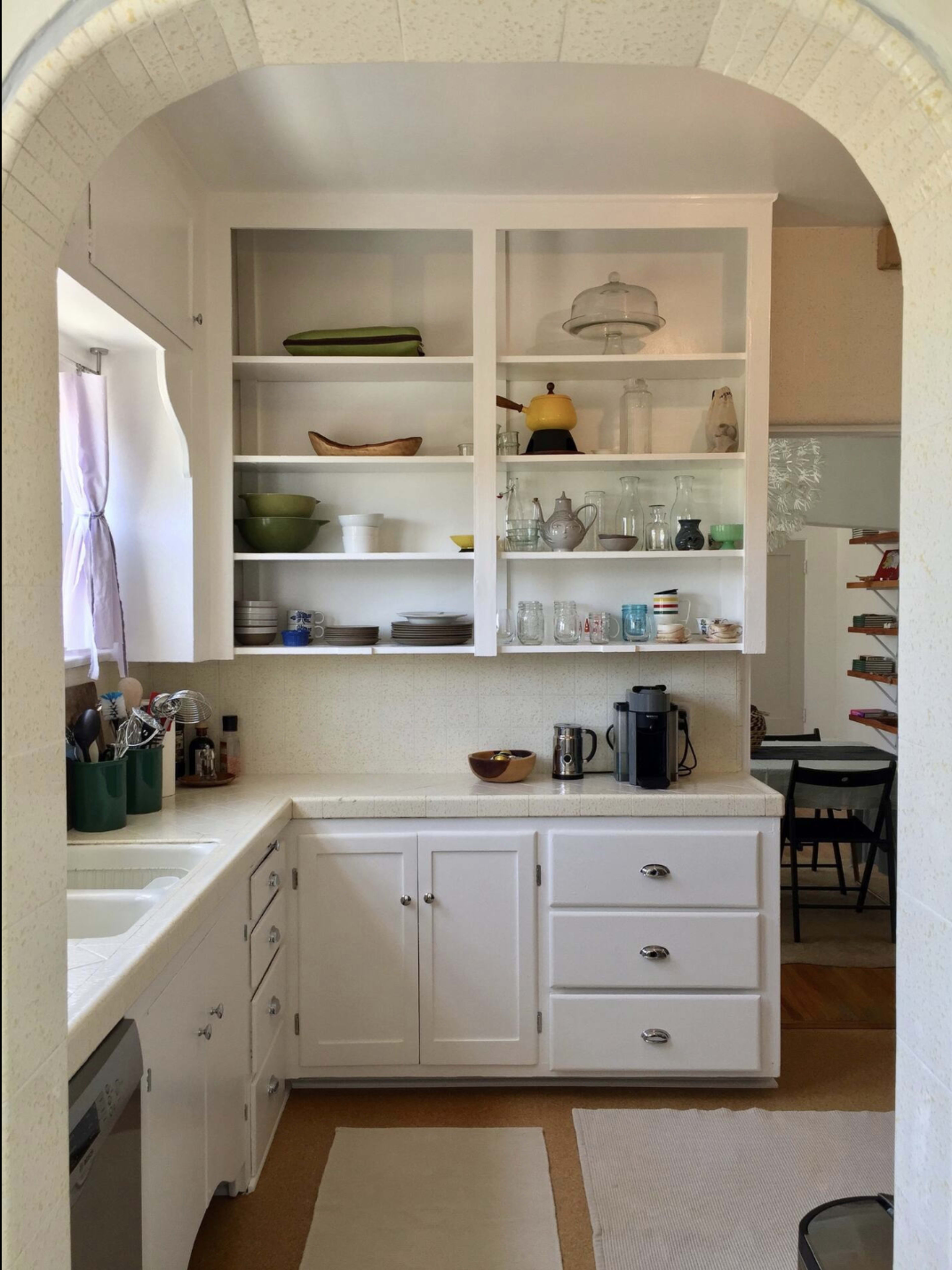 The image shows a tidy kitchen with white cabinets, open shelving displaying dishes and glassware, and a coffee maker on the counter.