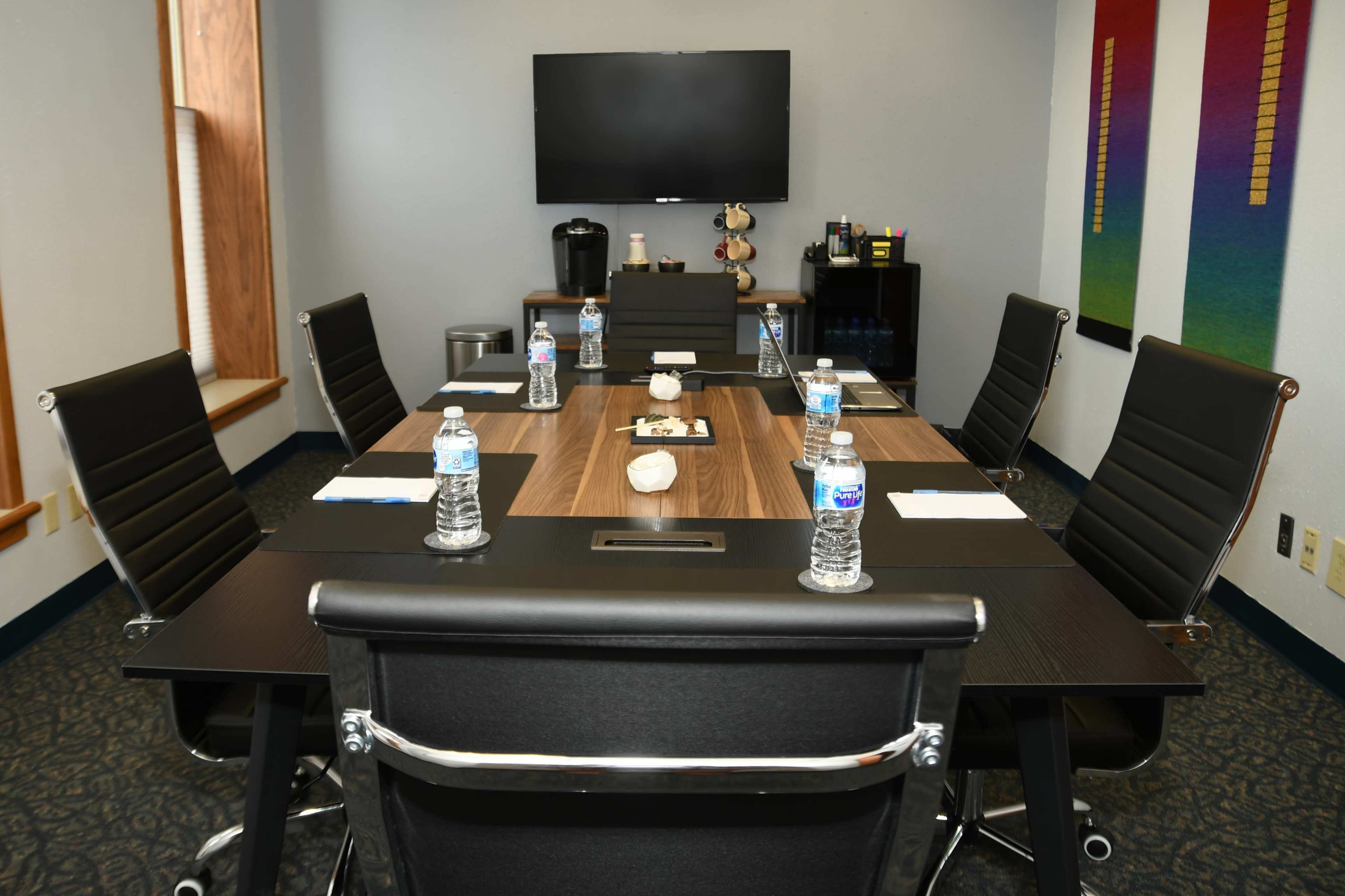 A conference room is set up with a long table surrounded by six chairs, each featuring a water bottle, utensils, and notepads, while a television is mounted on the wall.