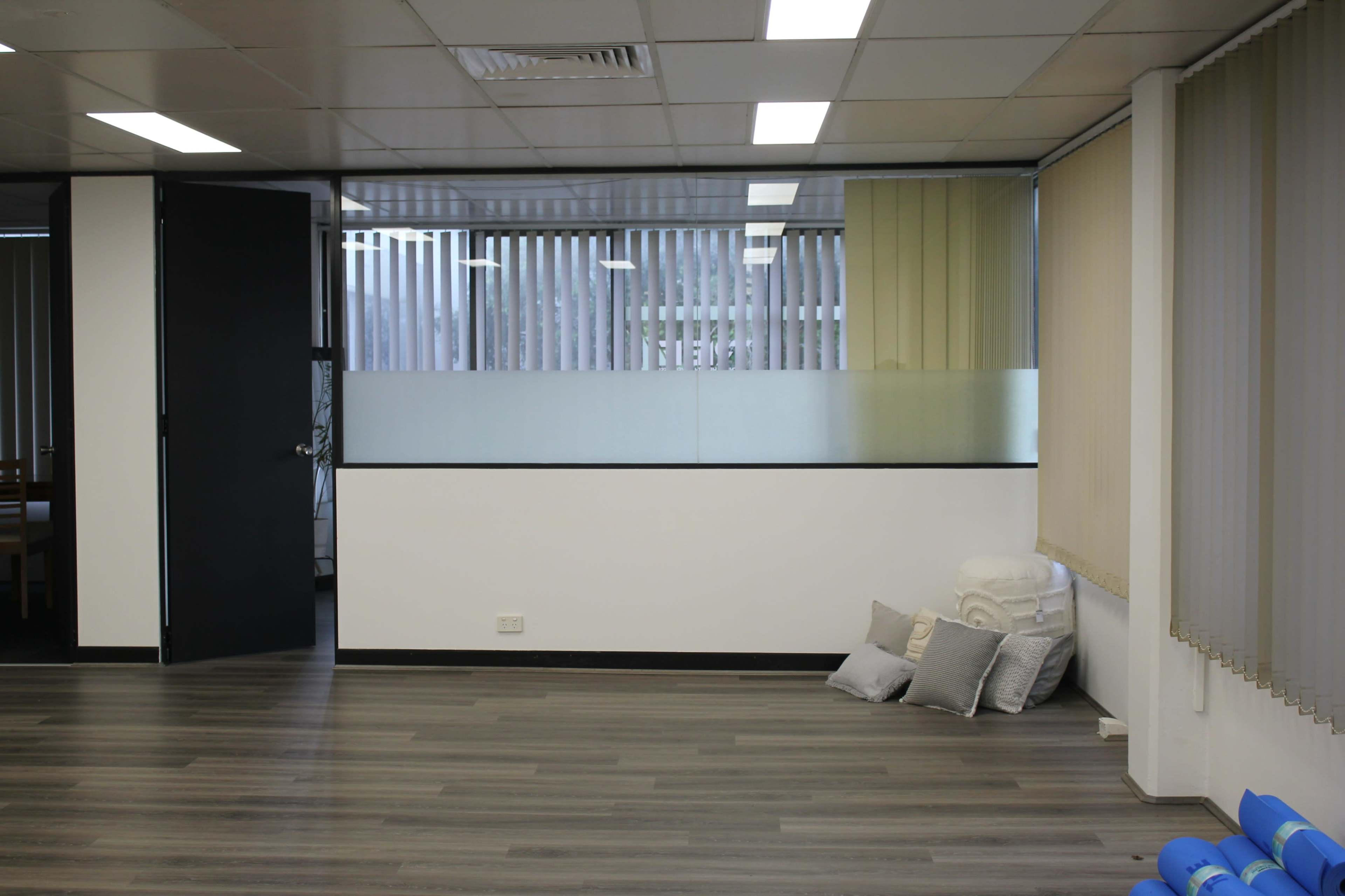 A modern office space with a doorway, a large window with vertical blinds, and neatly stacked cushions on the floor.