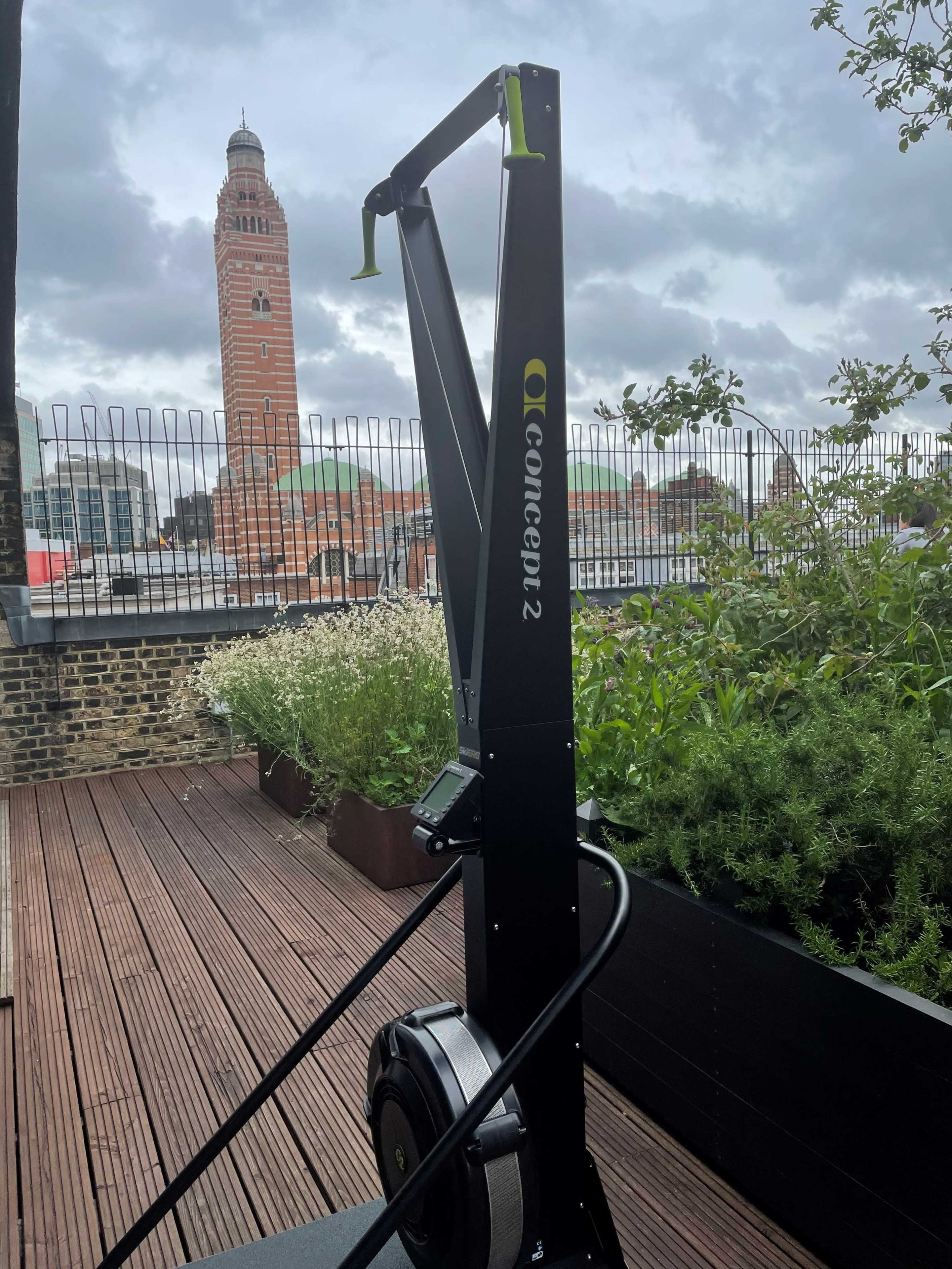 A Concept2 rowing machine is positioned on a wooden deck with a view of a tall clock tower in the background.