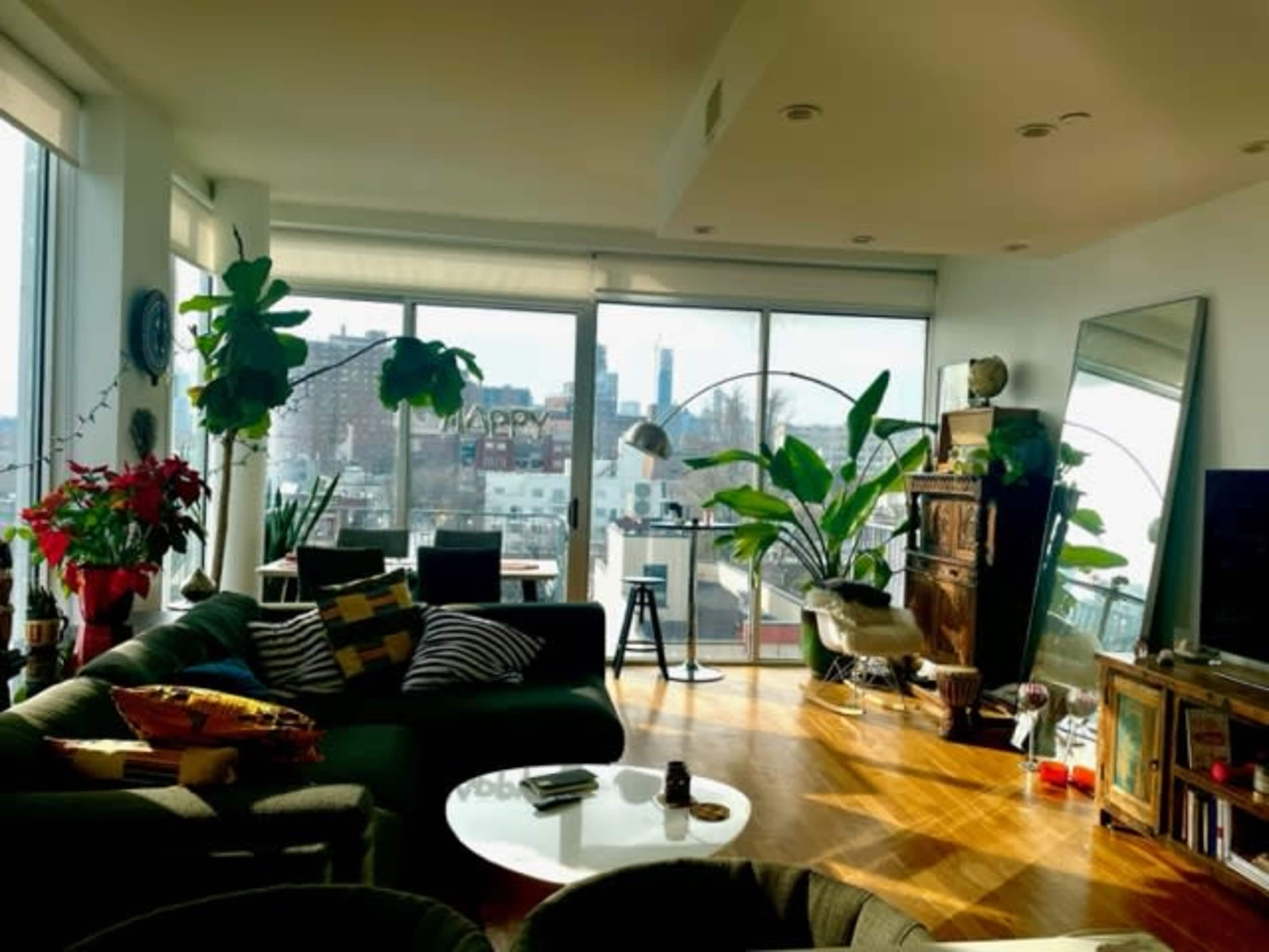 The image shows a modern living room with large windows, potted plants, and contemporary furniture overlooking a city skyline.
