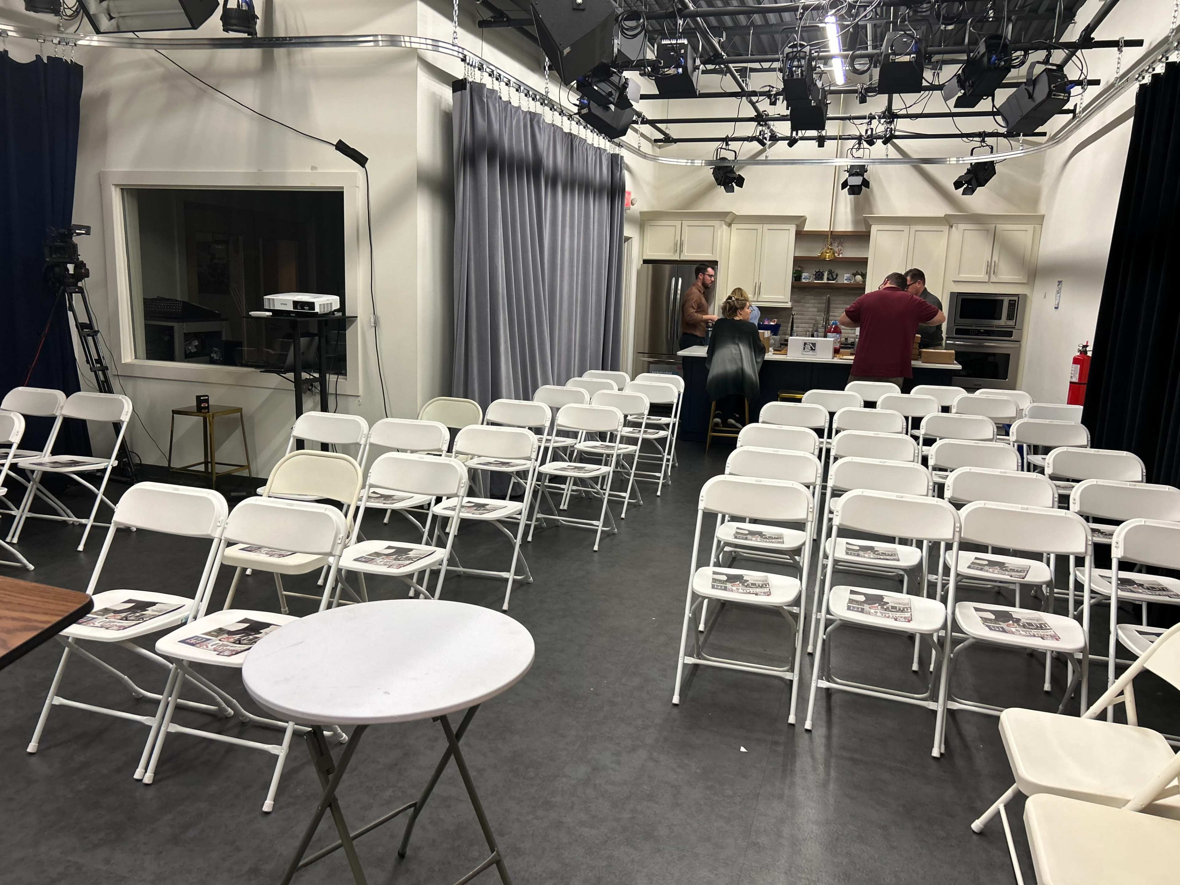 The room features a setup of white folding chairs arranged in rows, with a small kitchen area in the background and people preparing for an event.