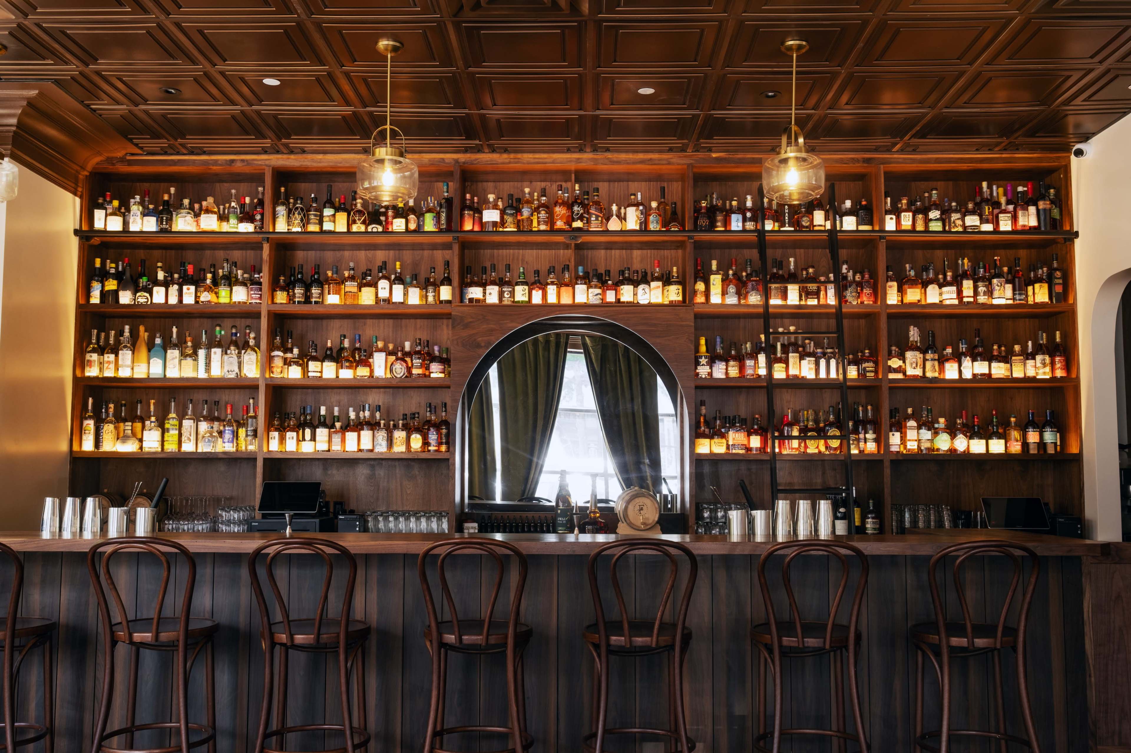 The image shows a wooden bar with a wall of illuminated shelves displaying a variety of liquor bottles, alongside a row of dark wooden stools.