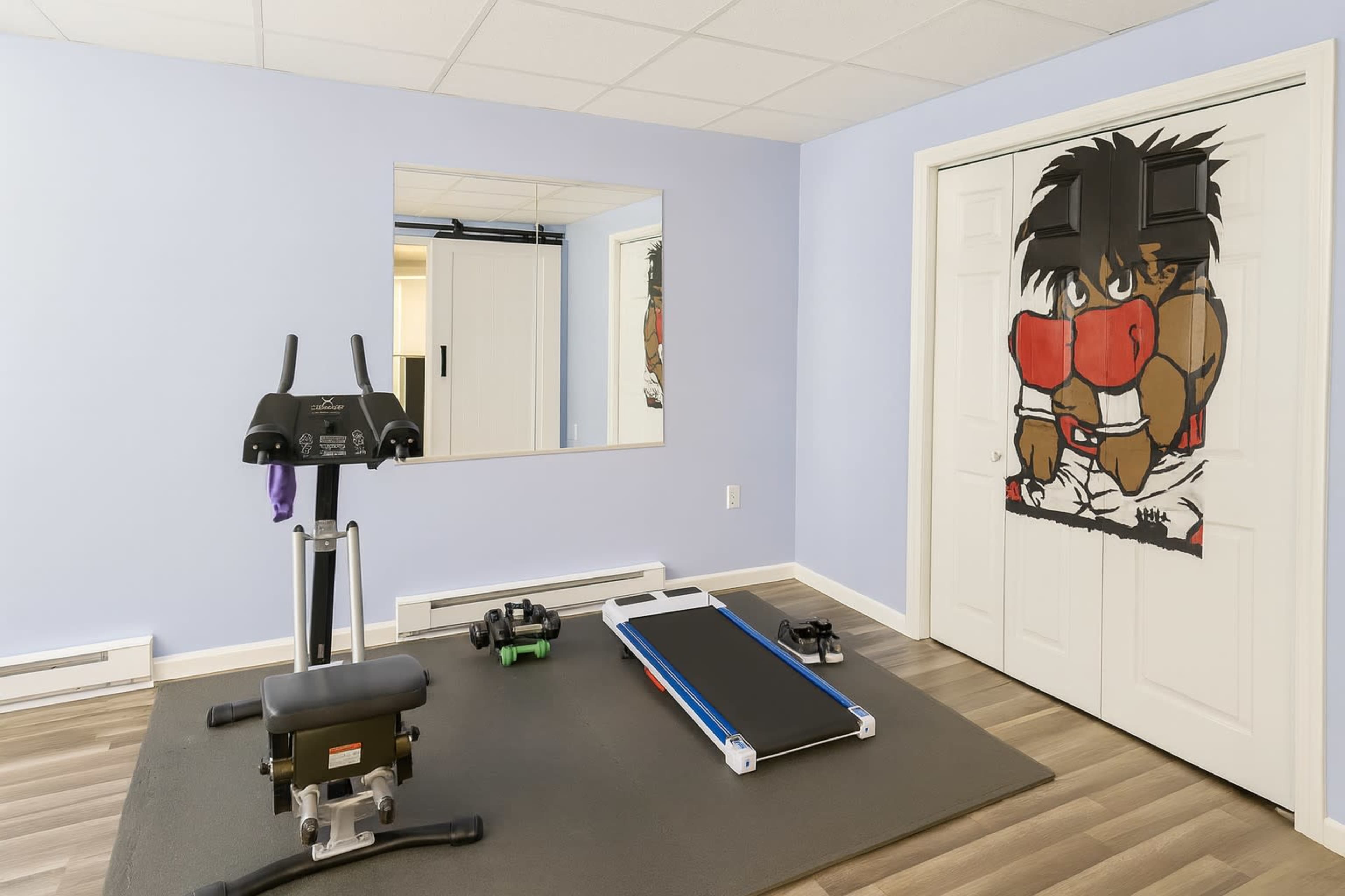 The image shows a home gym with exercise equipment, including a stationary bike, weights, and a treadmill, alongside a wall featuring a mural of a boxer.