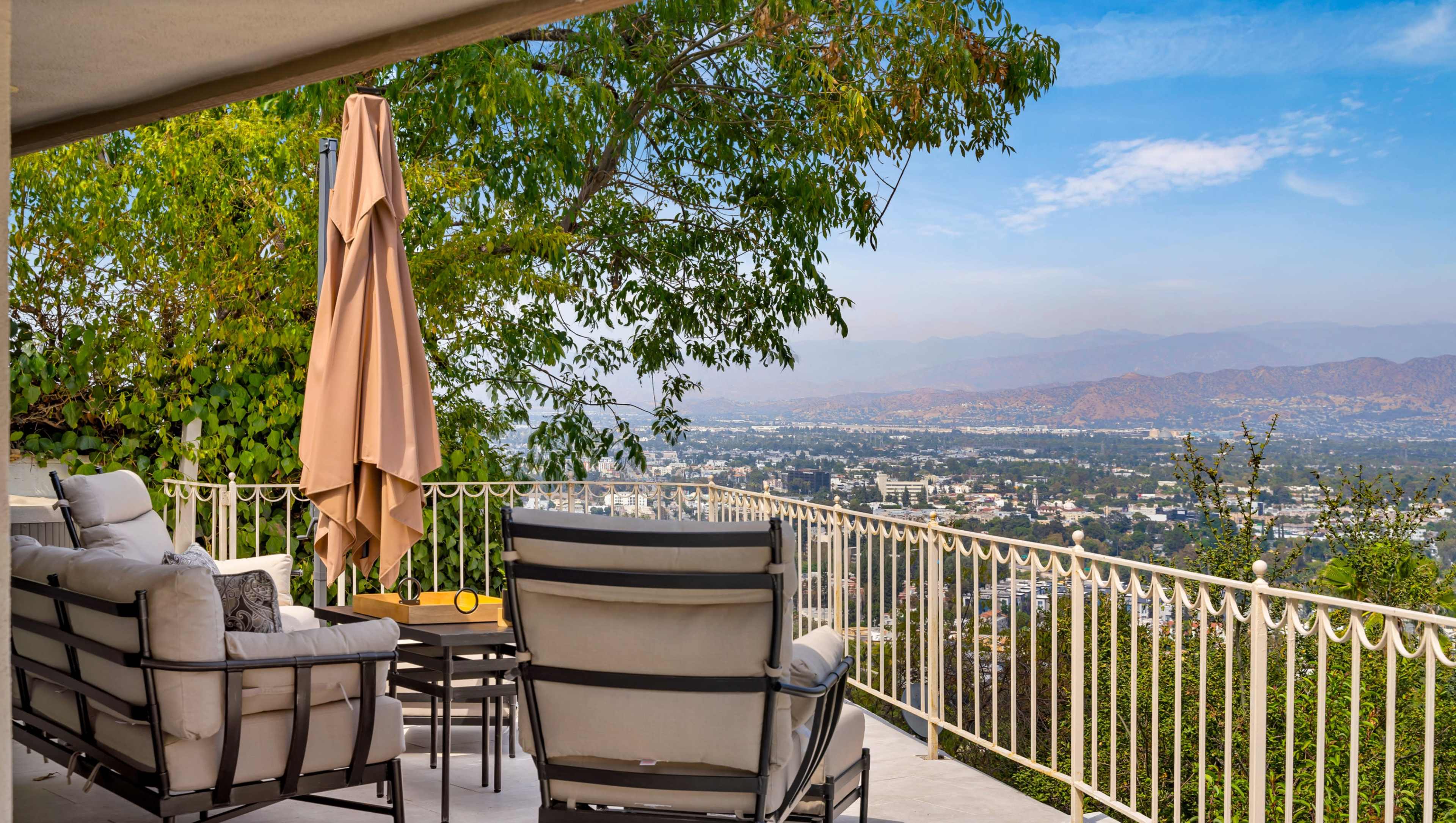 A patio area features outdoor furniture and a large umbrella, overlooking a sprawling valley with mountains in the distance.