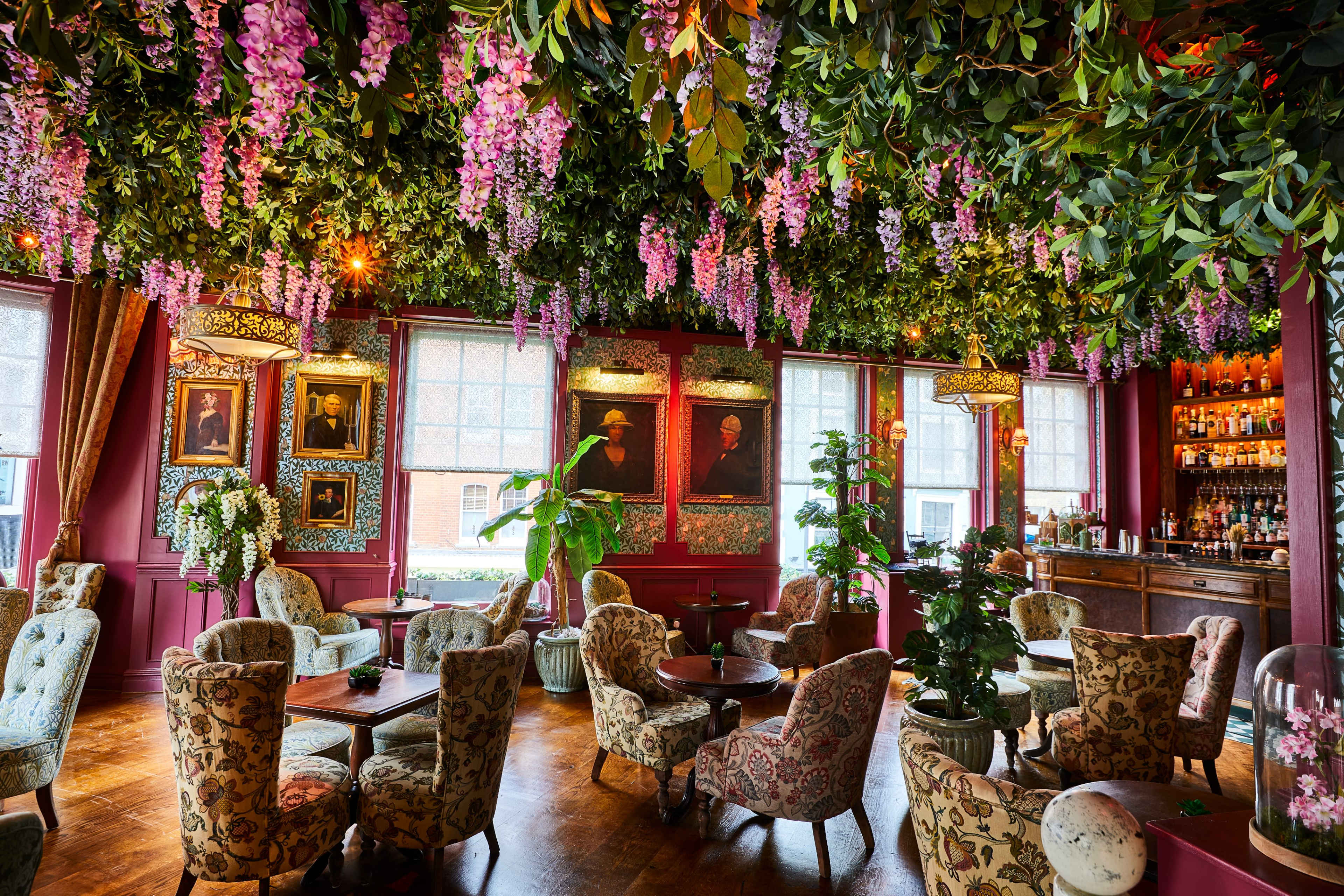 The image shows a cozy restaurant interior adorned with hanging flowers and greenery, featuring vintage portraits on the walls and elegantly patterned chairs around wooden tables.