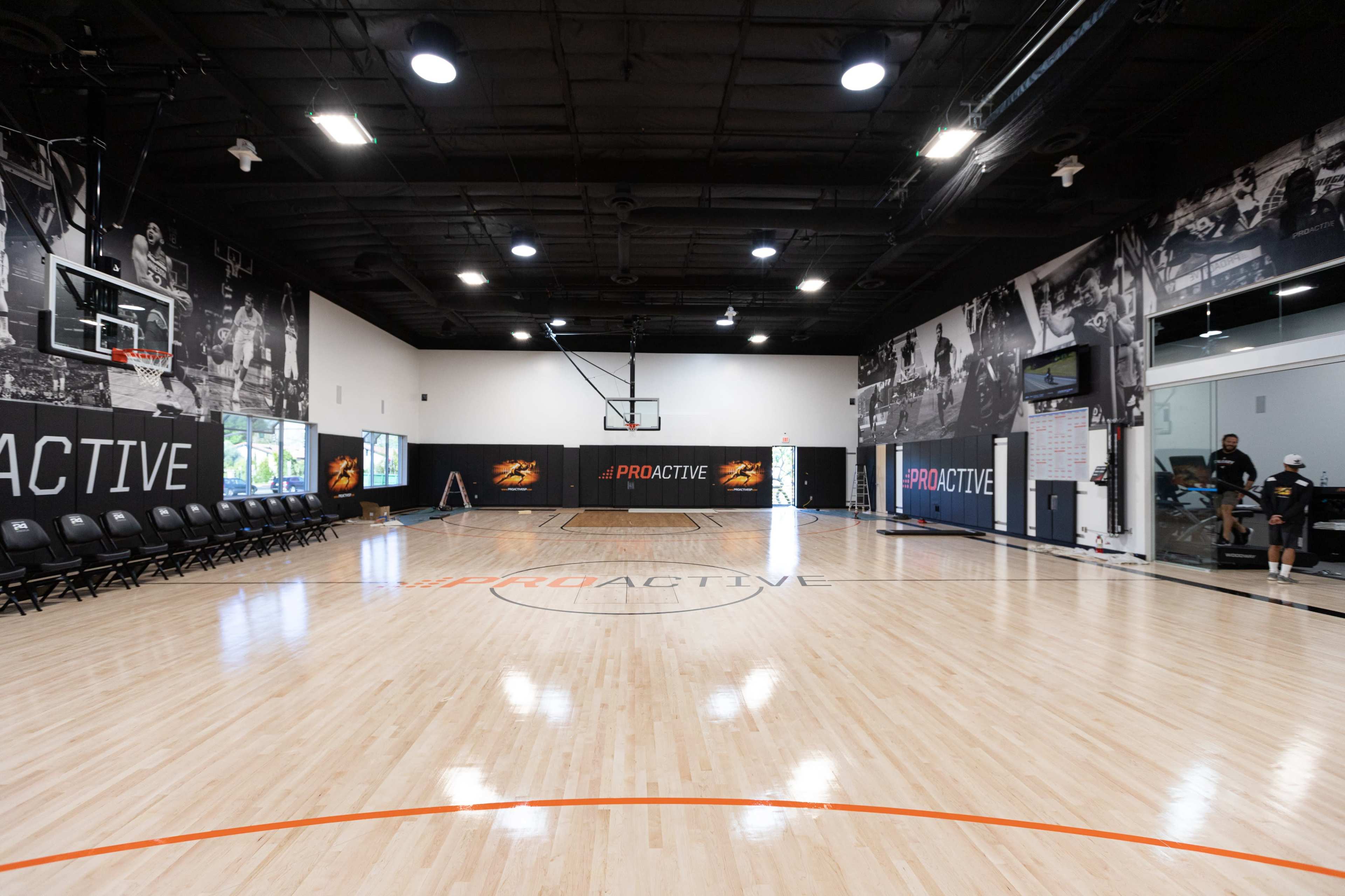 The image shows a basketball training facility with a polished wooden court, lined with black seating on one side and large murals on the walls.