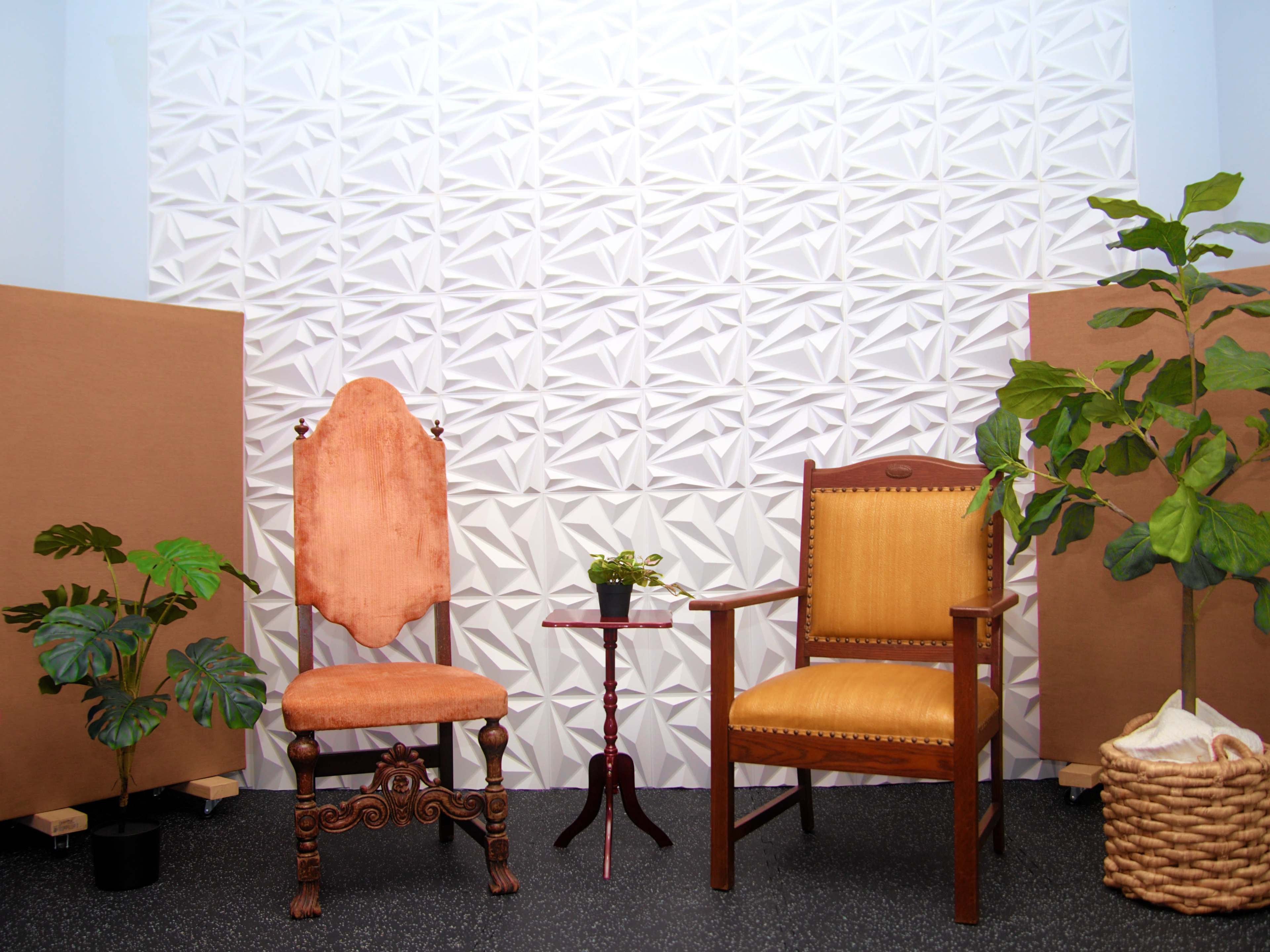 The image shows a small styled indoor corner featuring two chairs, a side table, a potted plant, and decorative panels in the background.