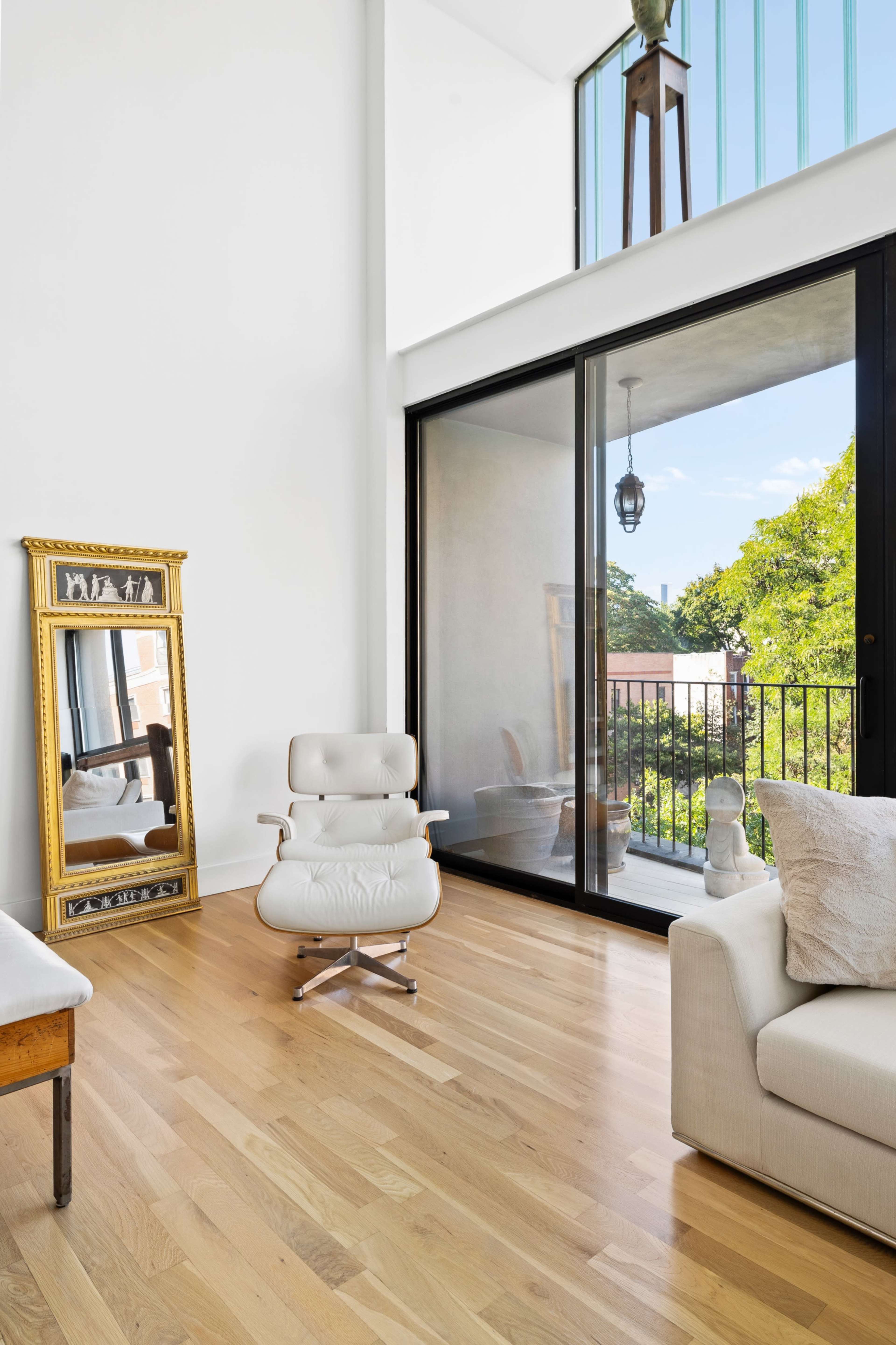 The image features a modern living room with a large glass door leading to a balcony, a white chair, a sofa, and a gold-framed mirror against a wall.
